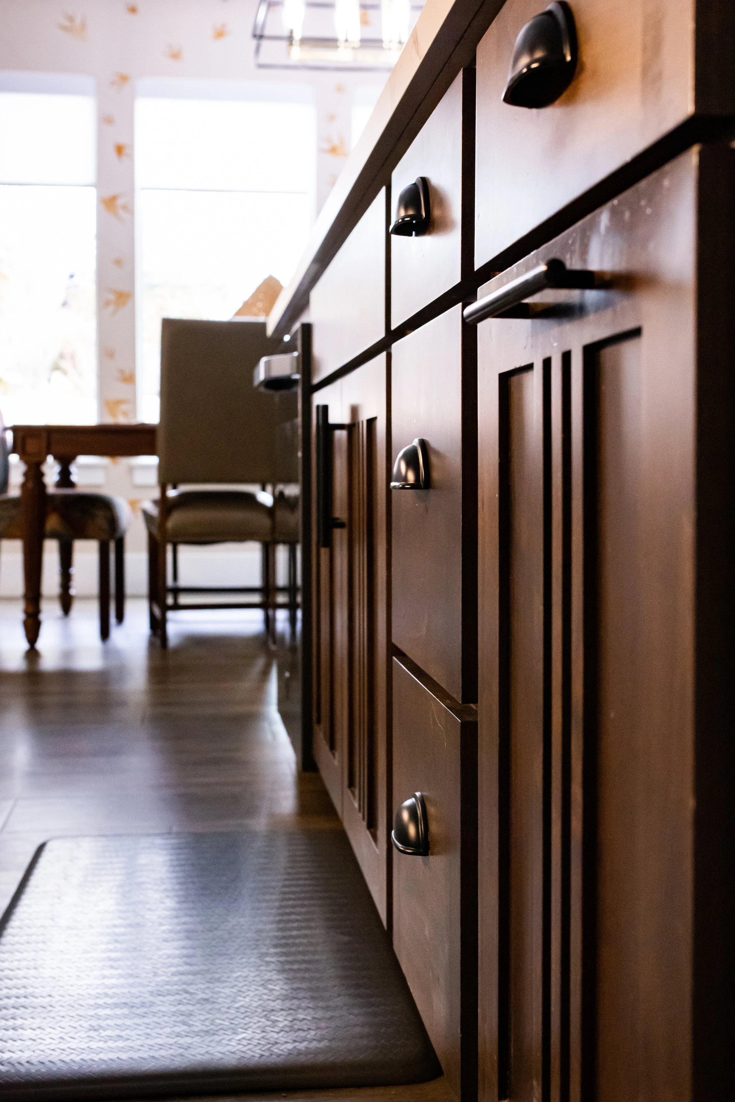 Close-up of a kitchen counter with wooden cabinets, silver handles, and a brown textured mat on the floor. In the background, a dining table with chairs and large windows letting in natural light.