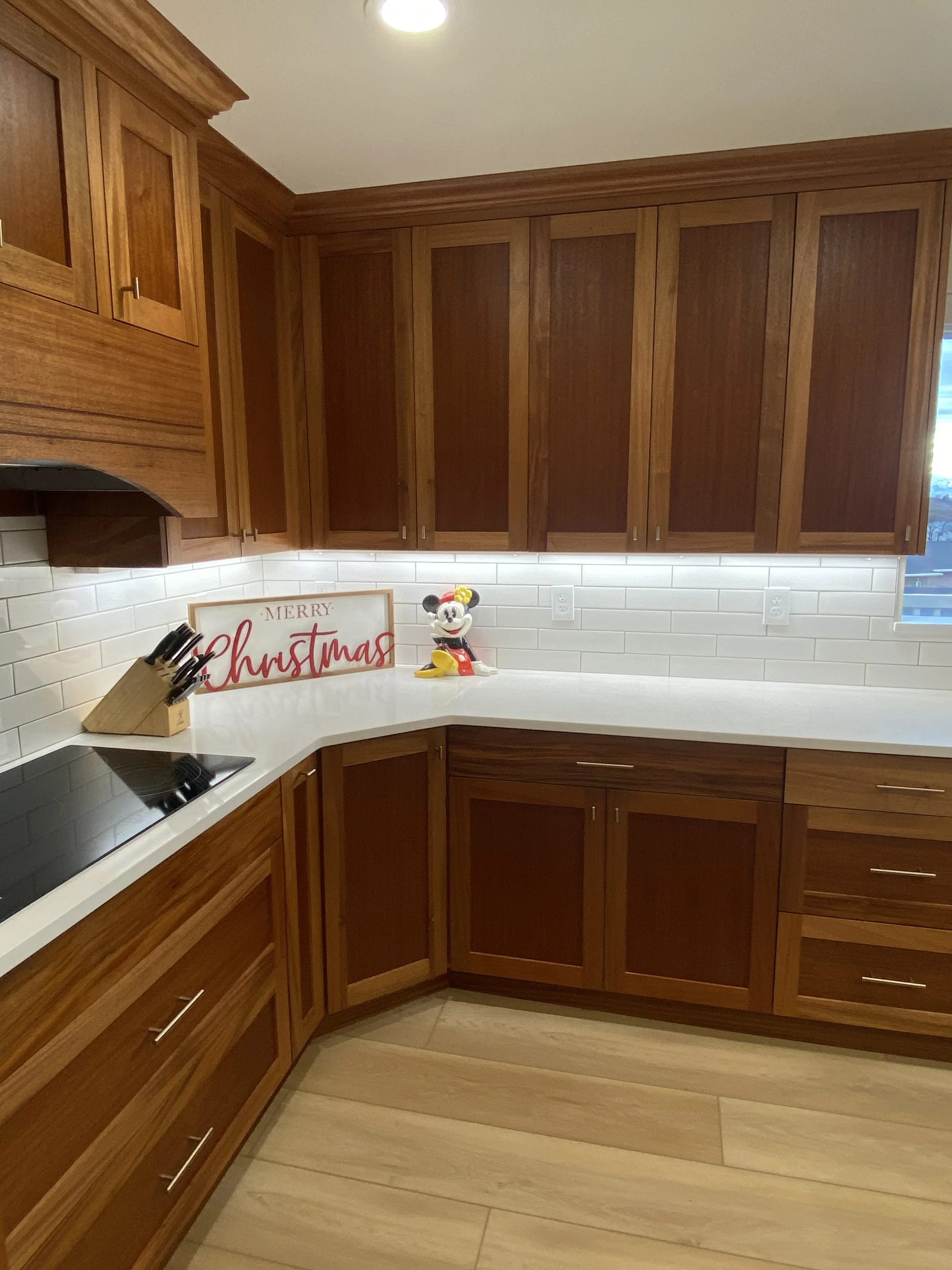 Wooden kitchen cabinets with white countertop, holiday decorations including a Mickey Mouse figure and a 'Merry Christmas' sign, and a window with natural light.