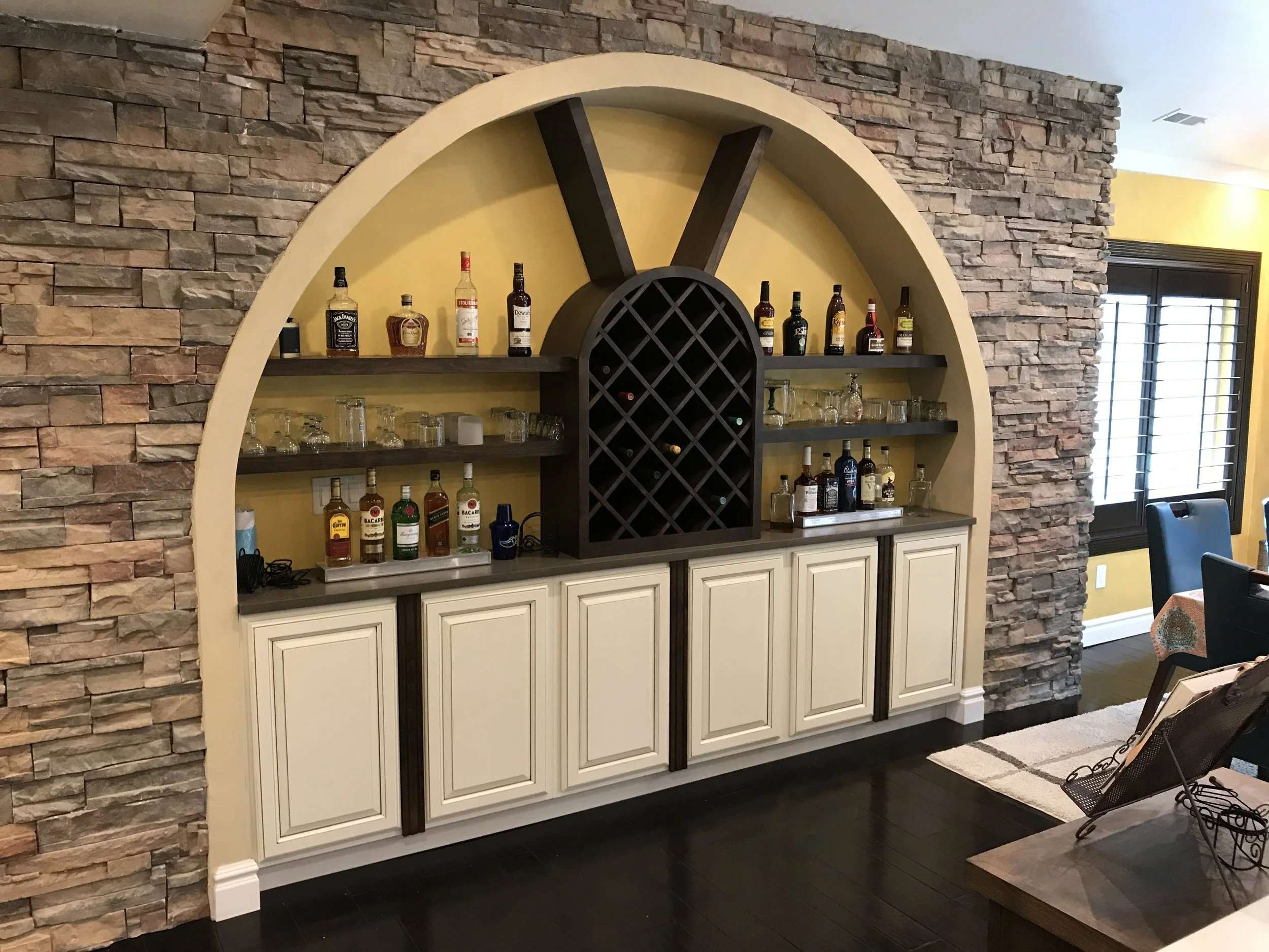Home bar with a wine rack on a yellow wall, surrounded by shelves with liquor bottles and glasses, and white cabinets below.