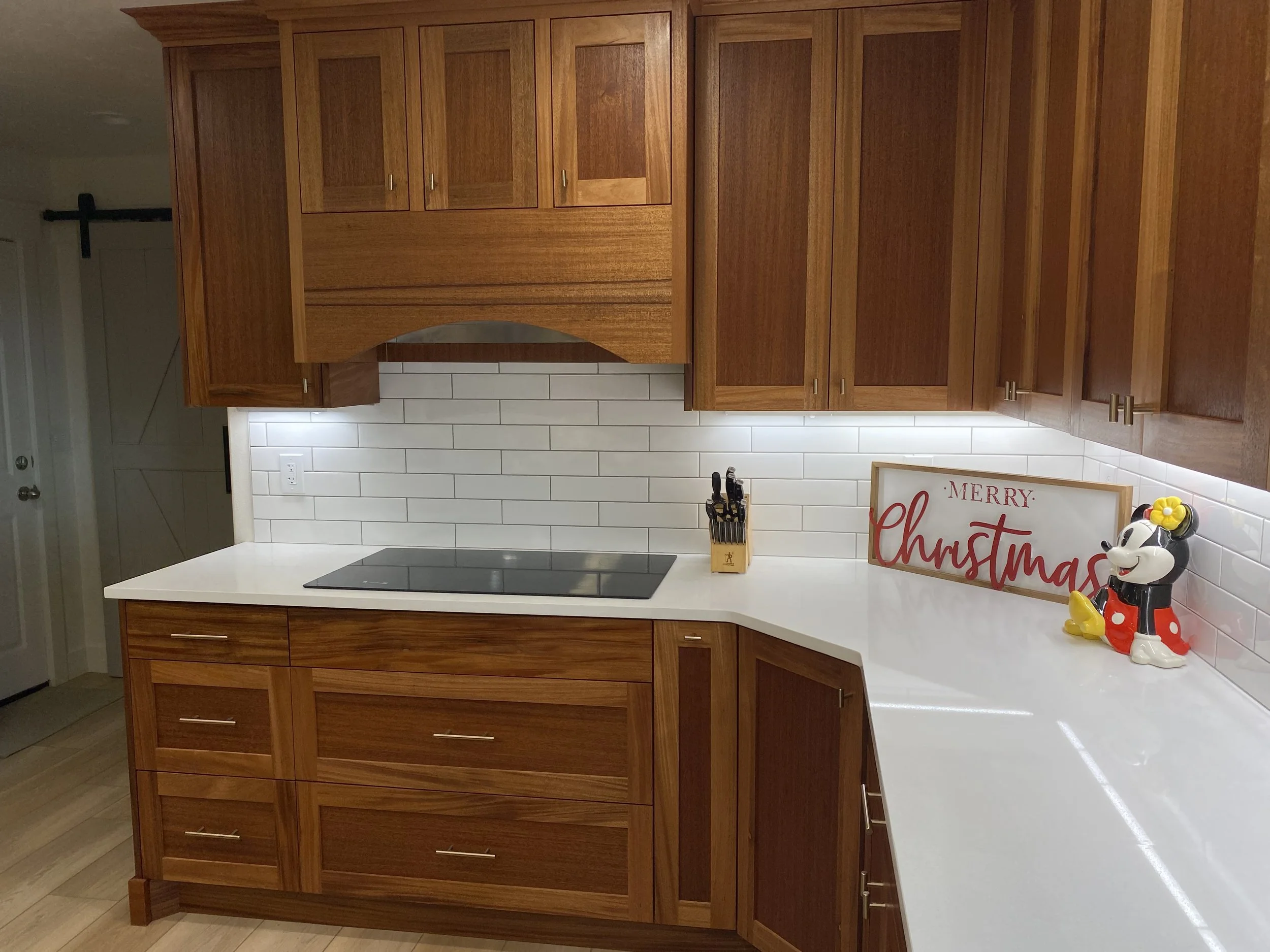 A kitchen with wooden cabinets, white countertops, and a white subway tile backsplash. There is a black cooktop, a set of knives in a wooden block, a 'Merry Christmas' sign, and a Minnie Mouse figurine on the counter. The kitchen has a modern, clean 