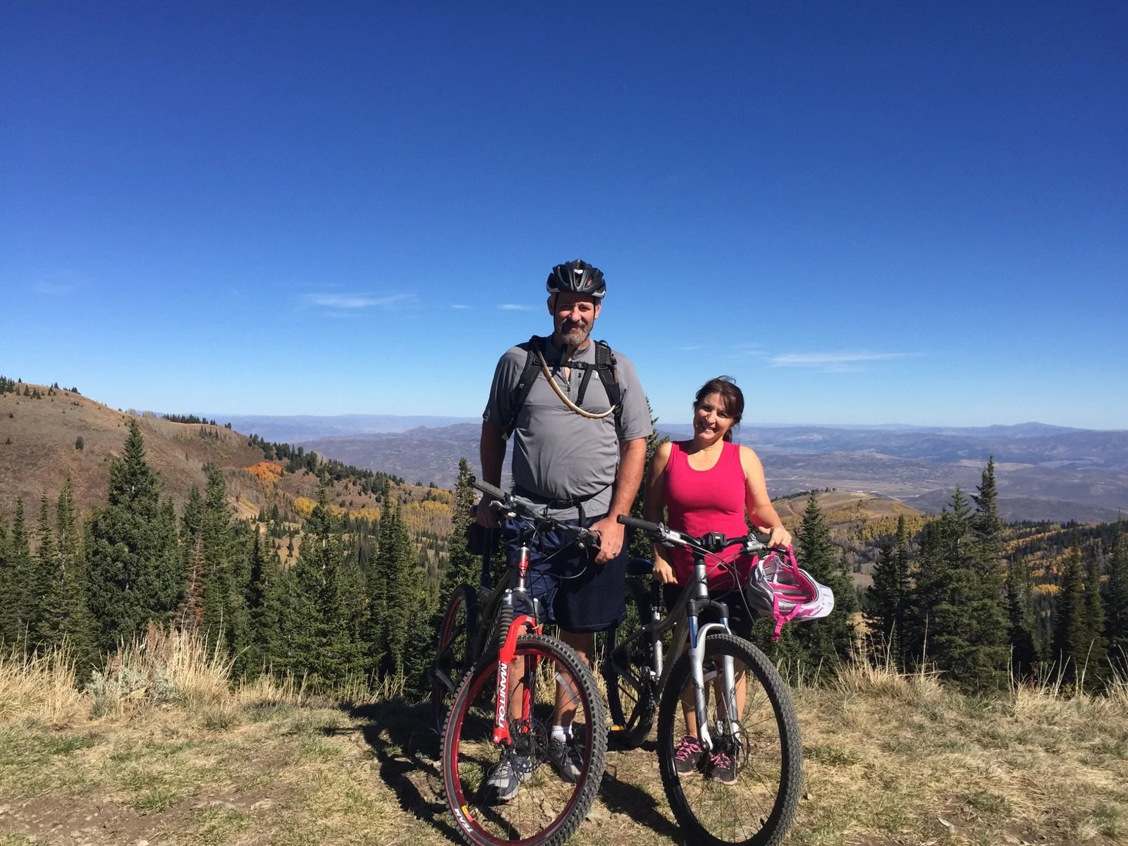 A man and a woman with bicycles standing on a mountain trail with a scenic view of forested mountains and valleys under a clear blue sky.