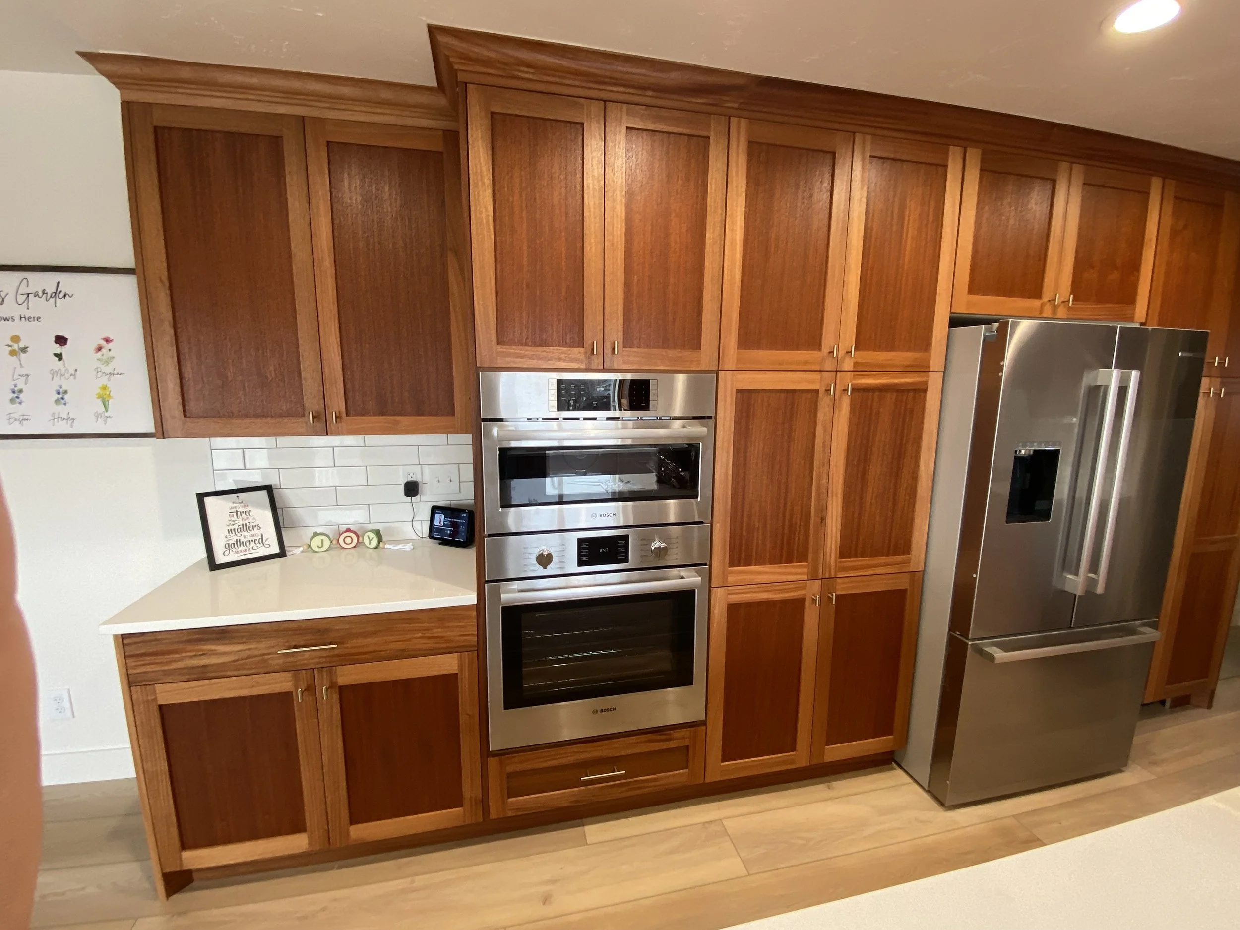 Kitchen with wooden cabinets, stainless steel oven and refrigerator, white backsplash, countertop with framed picture, small decorations, and a white wall with a colorful flower chart.