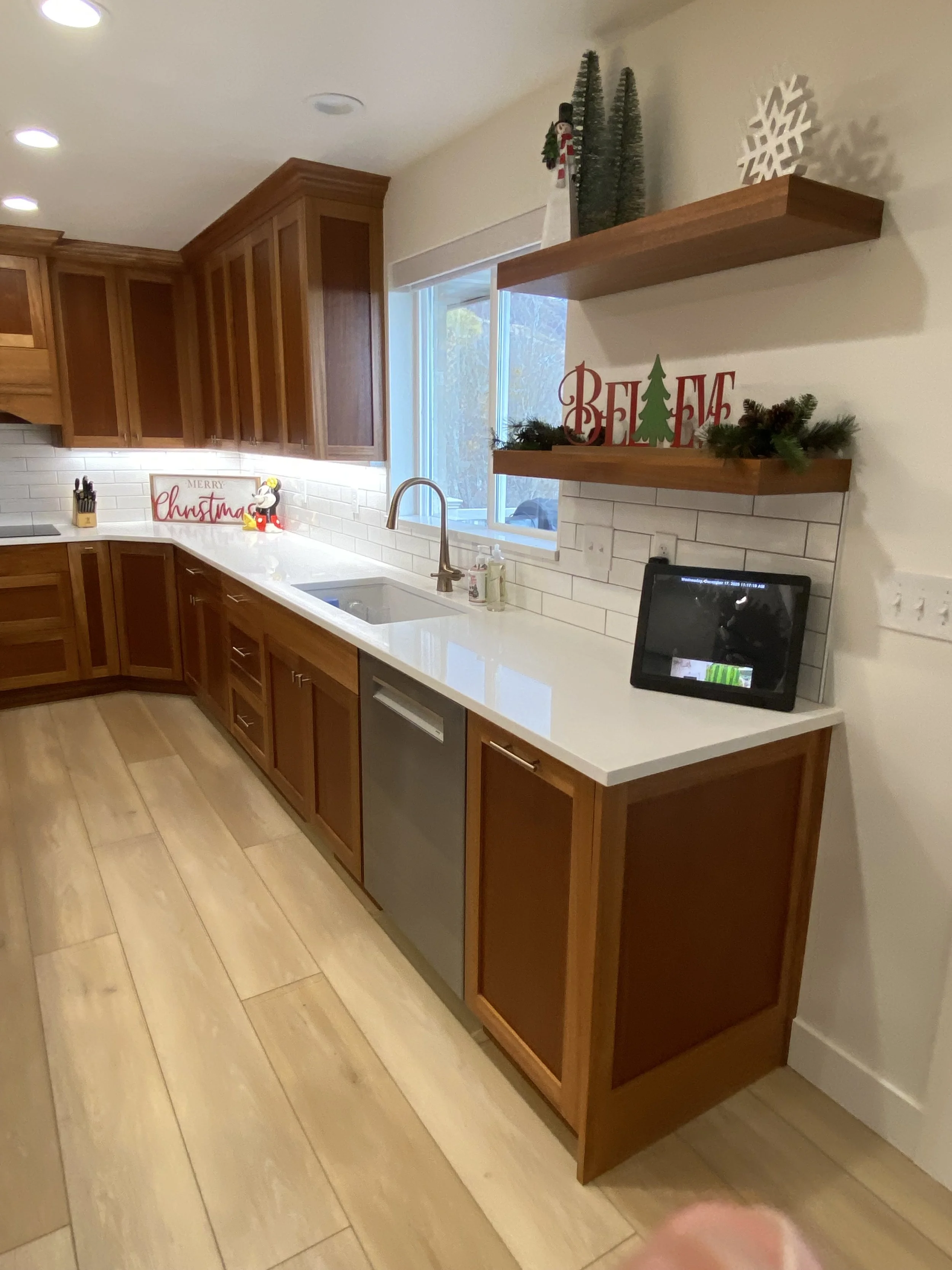 A kitchen with wooden cabinets, white countertop, and a sink. Christmas decorations include a sign that reads 'Merry Christmas,' a small Santa figurine, and holiday-themed ornaments on shelves and countertop.