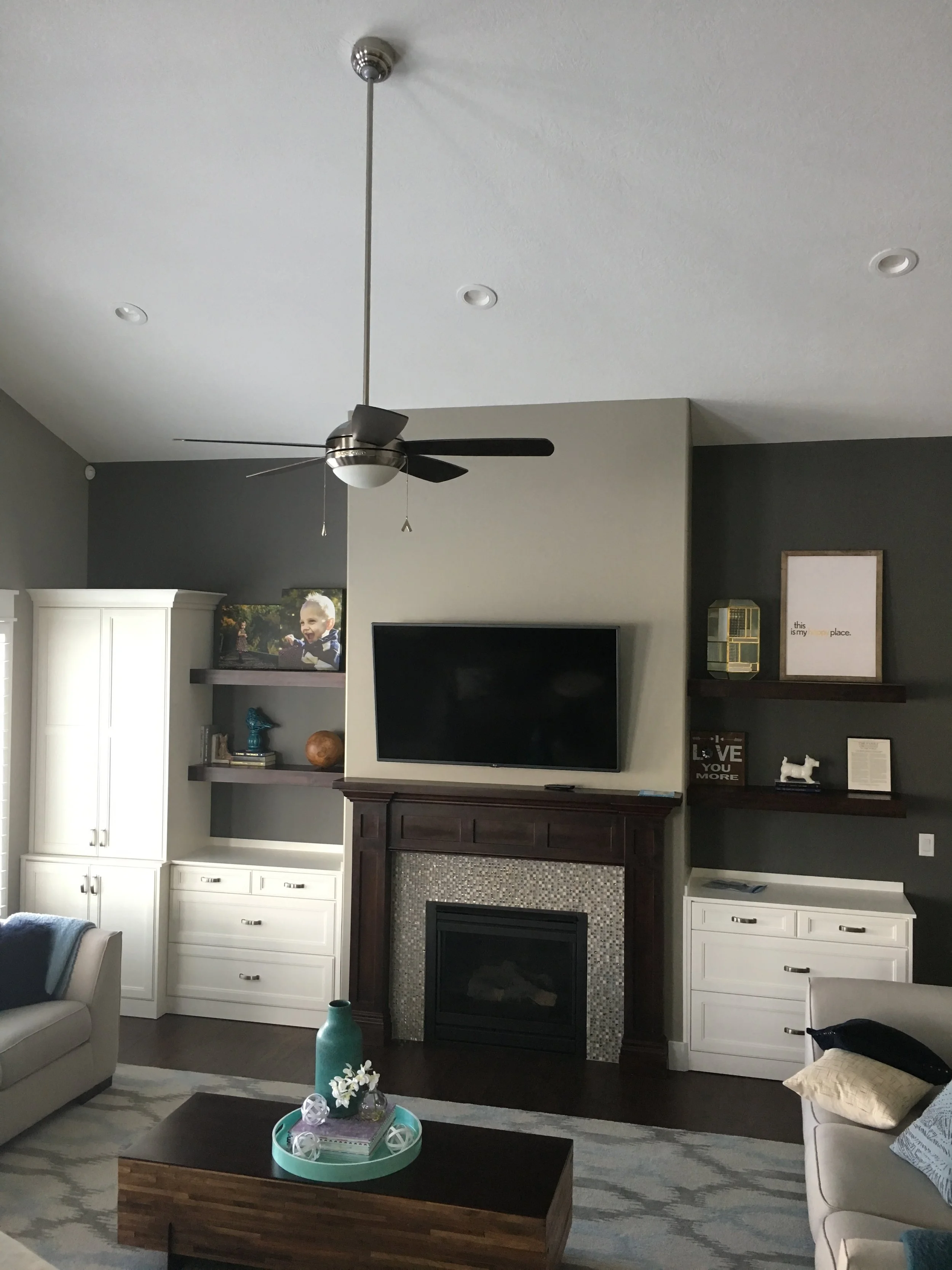 Living room with a ceiling fan, a fireplace with a TV above it, built-in white cabinets, dark wood shelves with decorative items, and a coffee table with a vase and decorative objects.