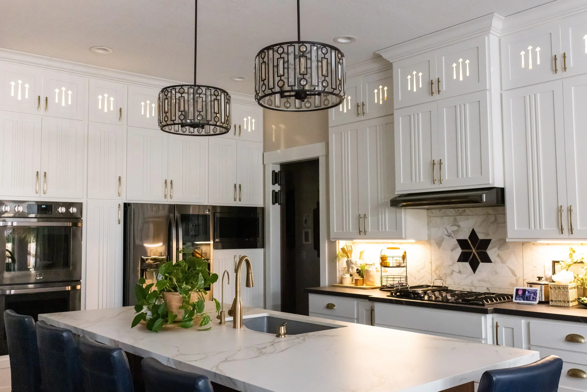 Modern kitchen with white cabinets, black and gold hardware, two black and gold pendant lights, a marble backsplash with a star design, marble kitchen island with a plant, gold faucet, stainless steel appliances, and a digital photo frame on the coun