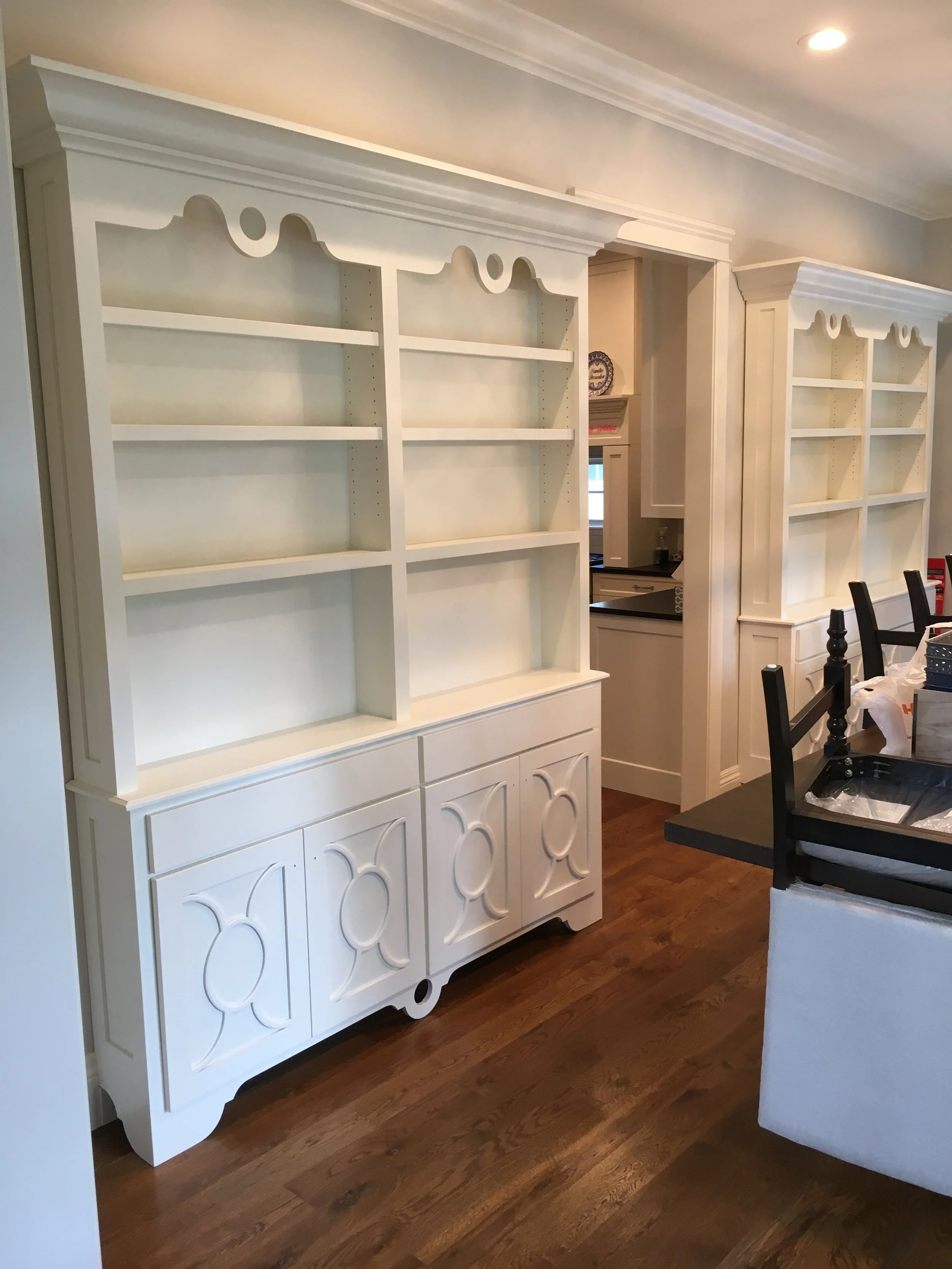 White wooden hutch with open shelves and decorative doors on the bottom, in a dining room with hardwood floor and a nearby chair and table.