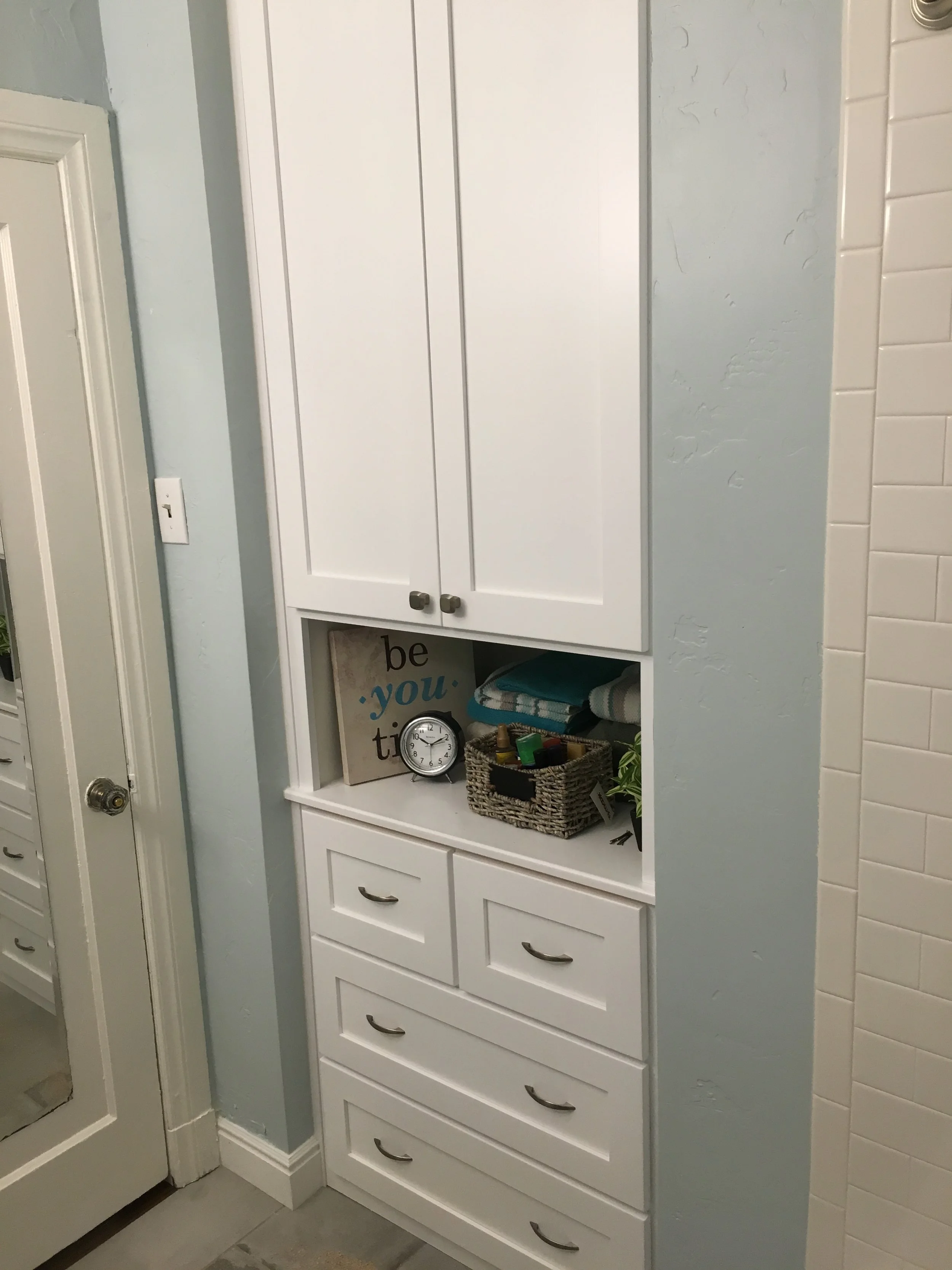 White kitchen cabinet with three large drawers at the bottom and closed doors at the top. An open shelf in the middle contains a small clock, a basket with small items, and some folded towels. A decorative sign reads 'be you tiful' behind the shelf, 