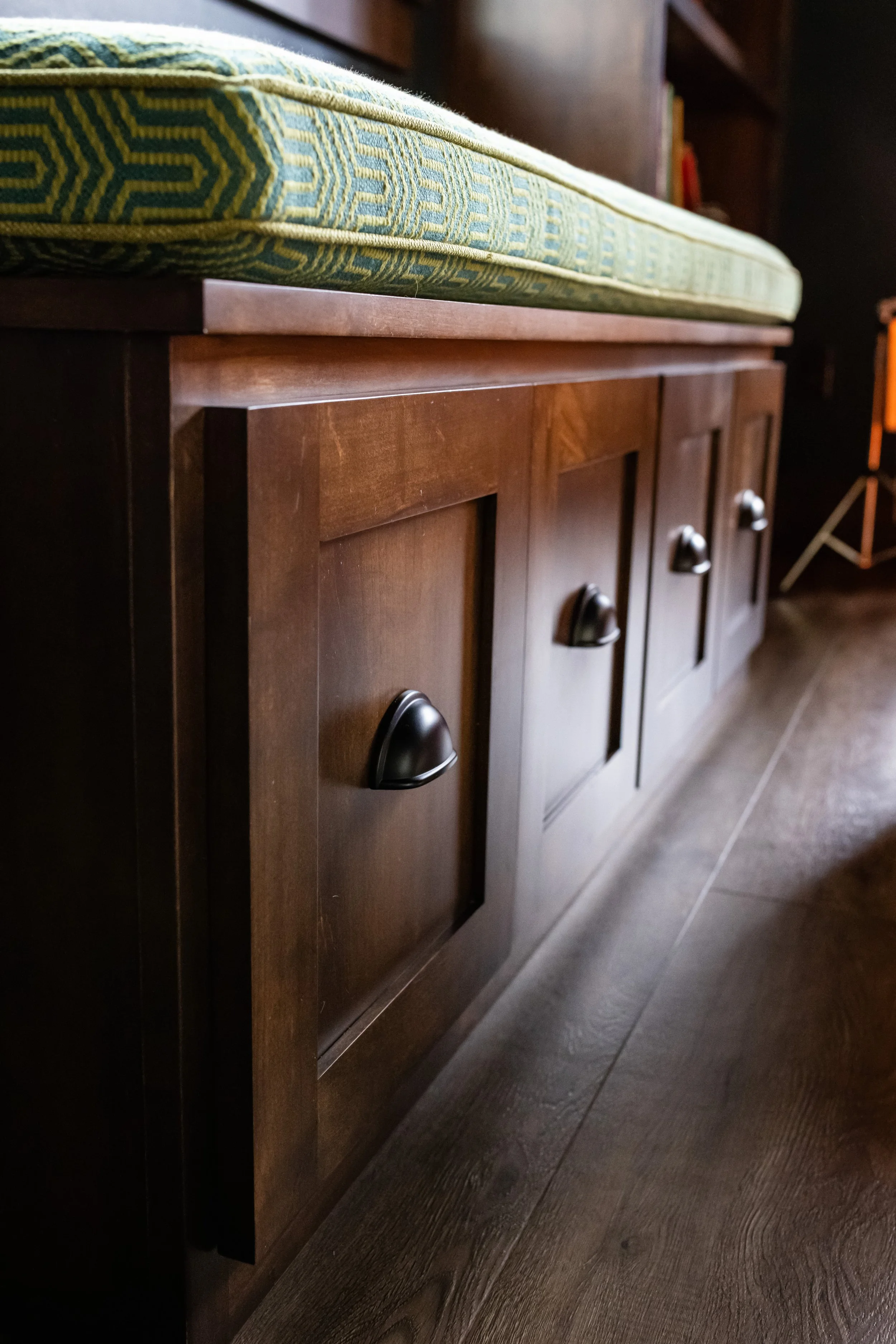 Close-up of a wooden cabinet with black handles and a cushioned top in a room with dark wood flooring and bookshelves.