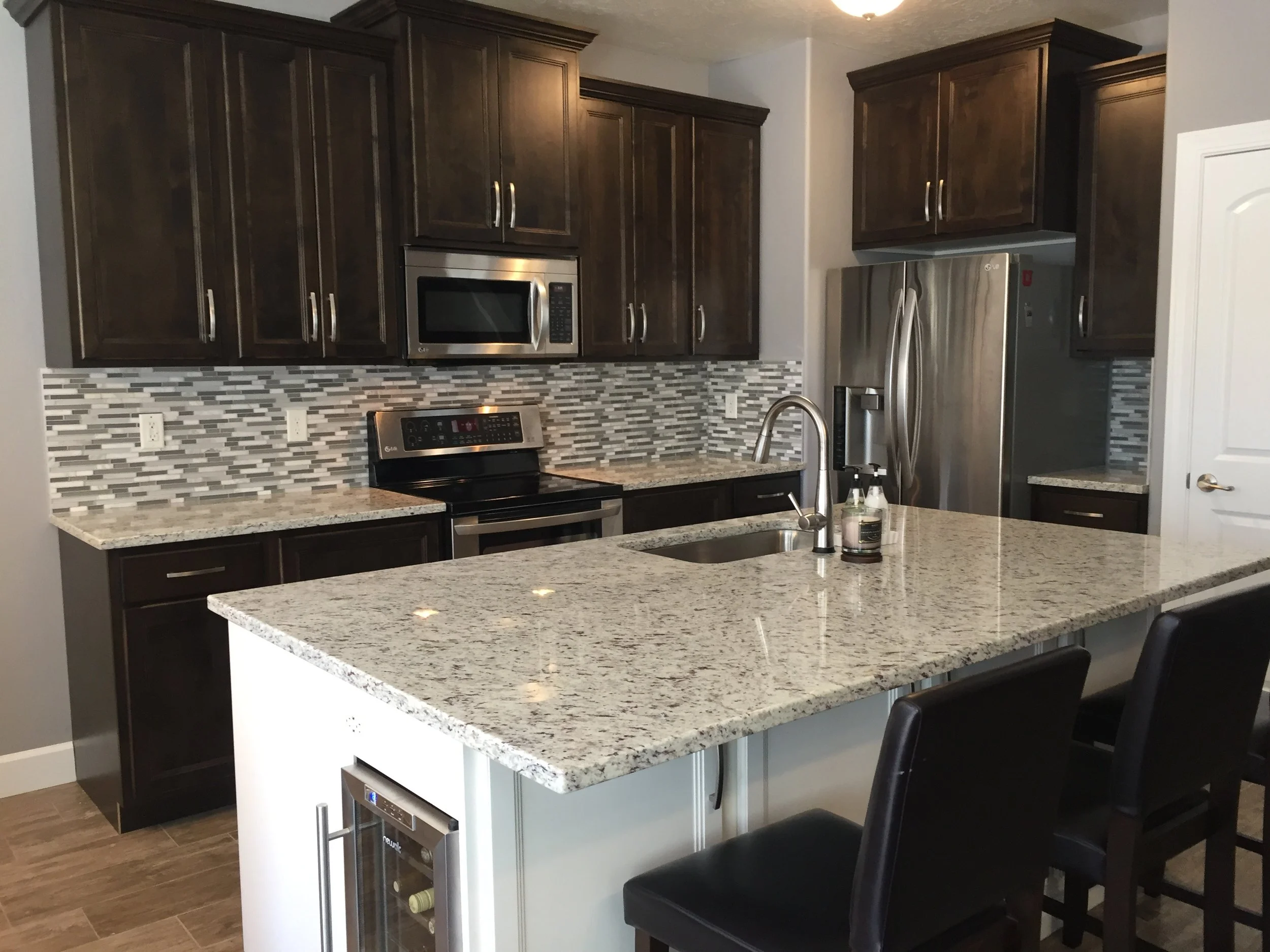 Modern kitchen with dark wooden cabinets, granite countertops, stainless steel appliances, mosaic tile backsplash, and a kitchen island with a sink and seating.