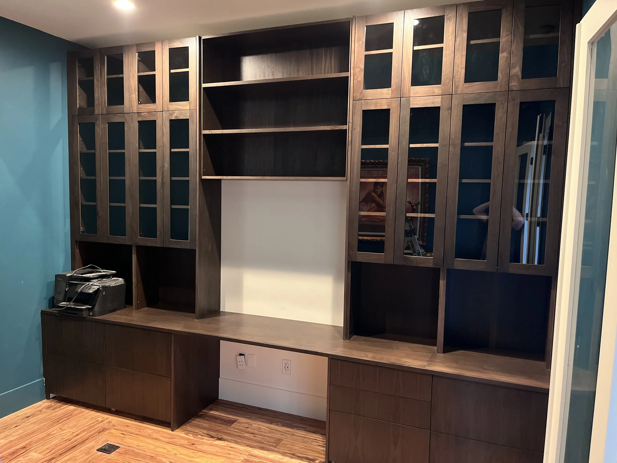A large wooden wall unit with glass doors, open shelves, and closed cabinets, with a printer on the left side and a white wall in the center.
