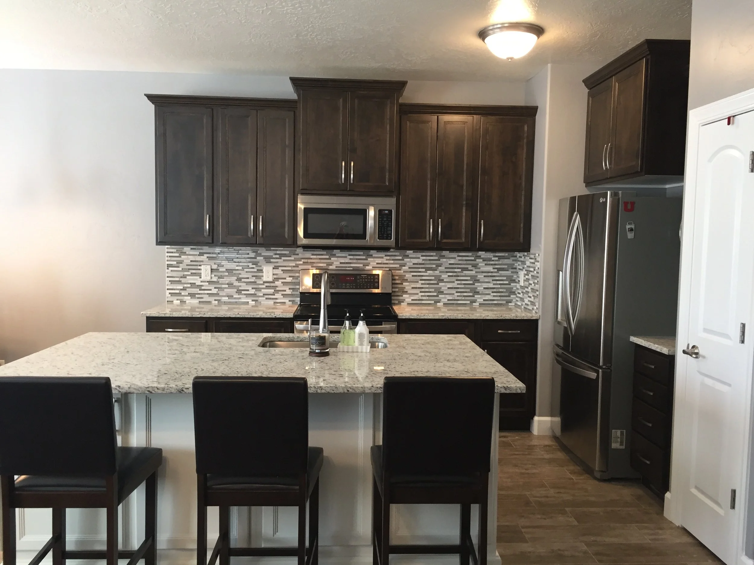 Modern kitchen with dark wood cabinets, a granite island with three black chairs, stainless steel appliances, a mosaic tile backsplash, and a white wall.