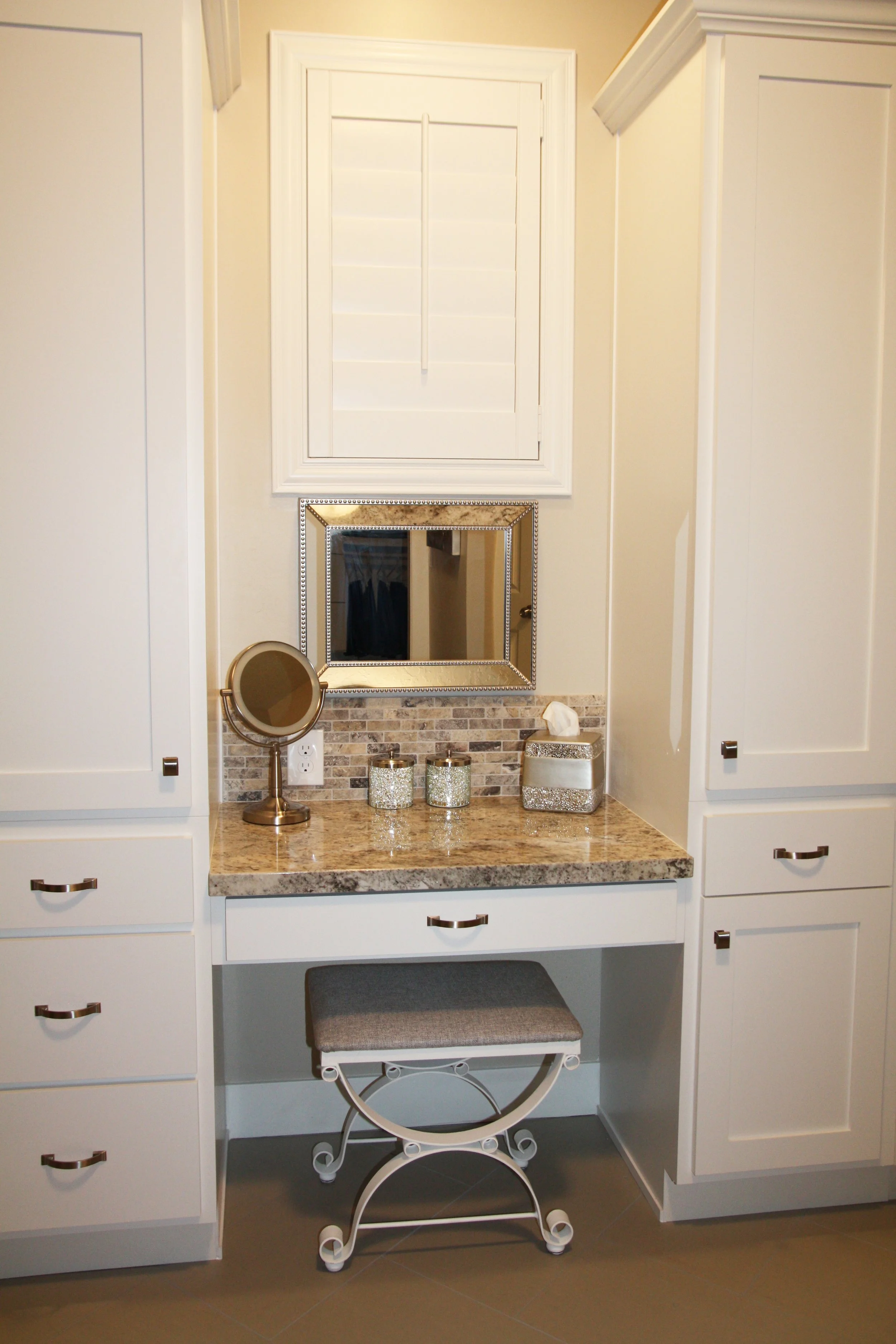 Bathroom vanity with white cabinets, a granite countertop, and a small vanity mirror. There are two decorative jars, a tissue box, and a small mirror on the countertop. A window with white shutters is above the vanity.