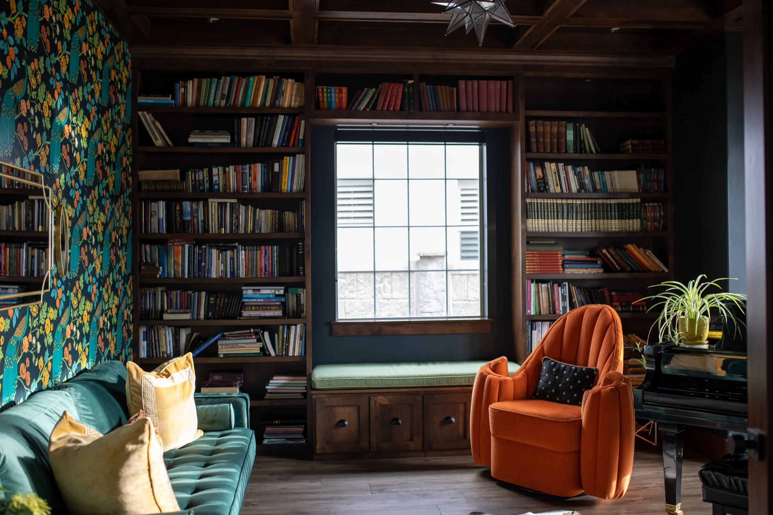 A cozy living room with a large window, dark wooden bookshelves filled with books, a green upholstered sofa with yellow cushions, an orange armchair with a black star-patterned pillow, a black piano with a potted plant on top, and a decorative star-s