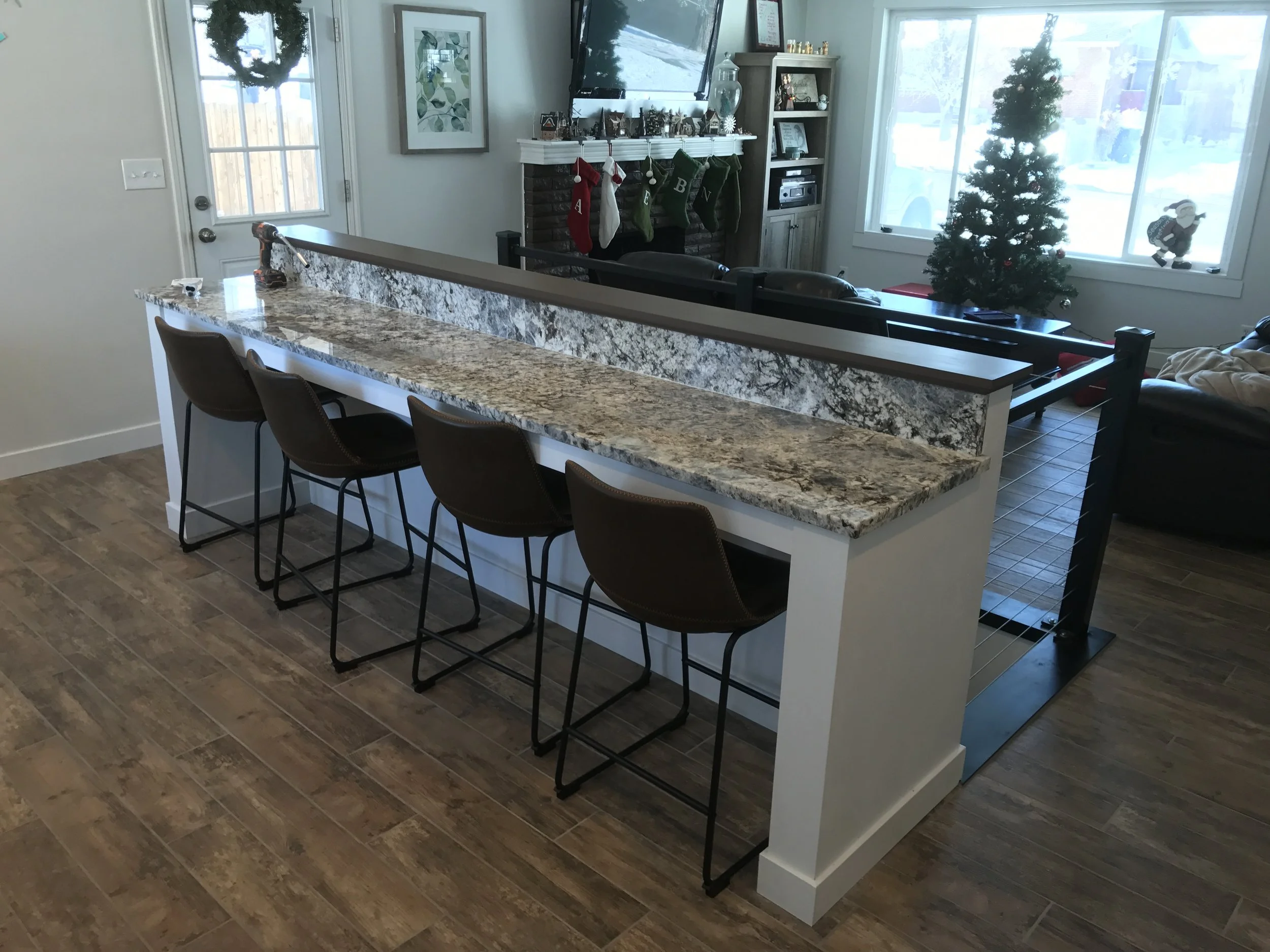 Kitchen island with three brown chairs, granite countertop, and a partial view of the living room with Christmas decorations in a window and a Christmas tree.