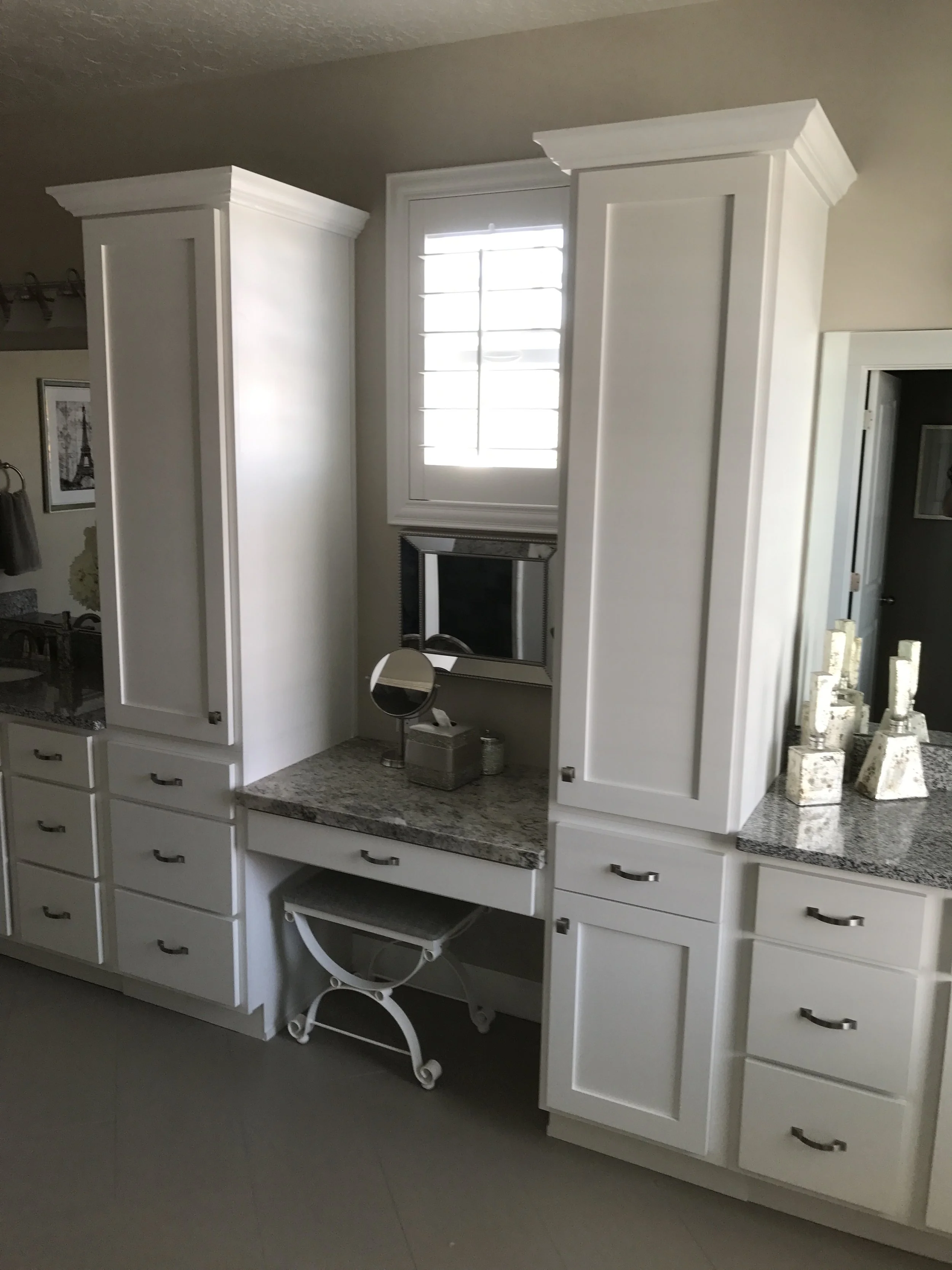 White built-in cabinets and drawers with granite countertops, a small vanity with a mirror, a window with white shutters, and some decorative items on the countertops.