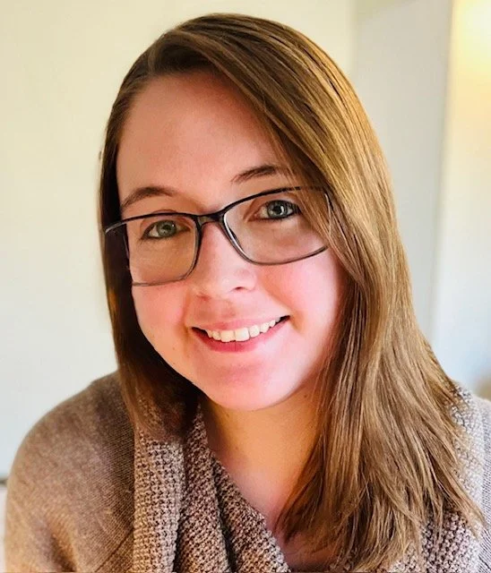 Close-up of a smiling woman with long brown hair, glasses, and wearing a brown sweater.