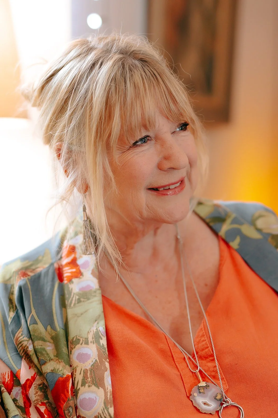 Close-up of a smiling older woman with red hair, wearing an orange top, a floral-patterned jacket, and a necklace with a large pendant. She is indoors, with warm lighting in the background.
