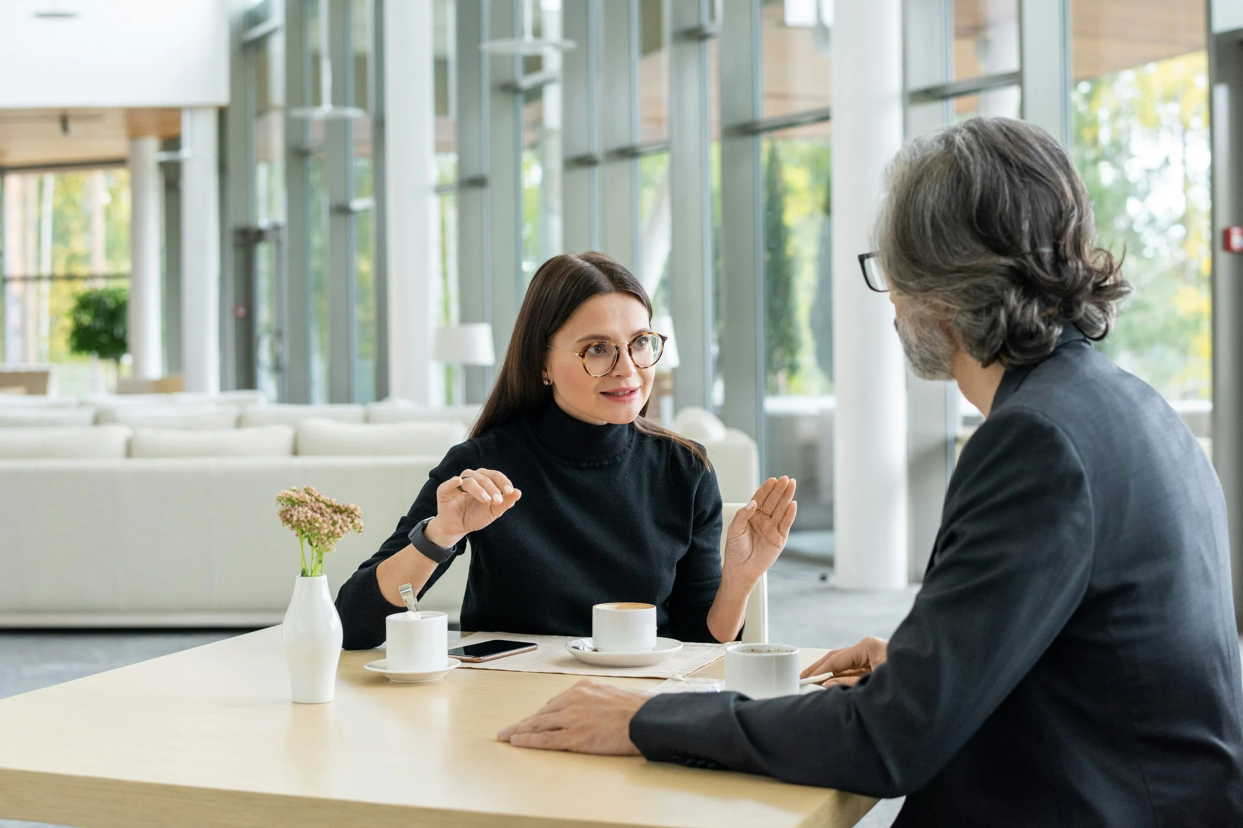 A therapist and client sit in a light-filled room talking face to face. THey are drinking coffee.