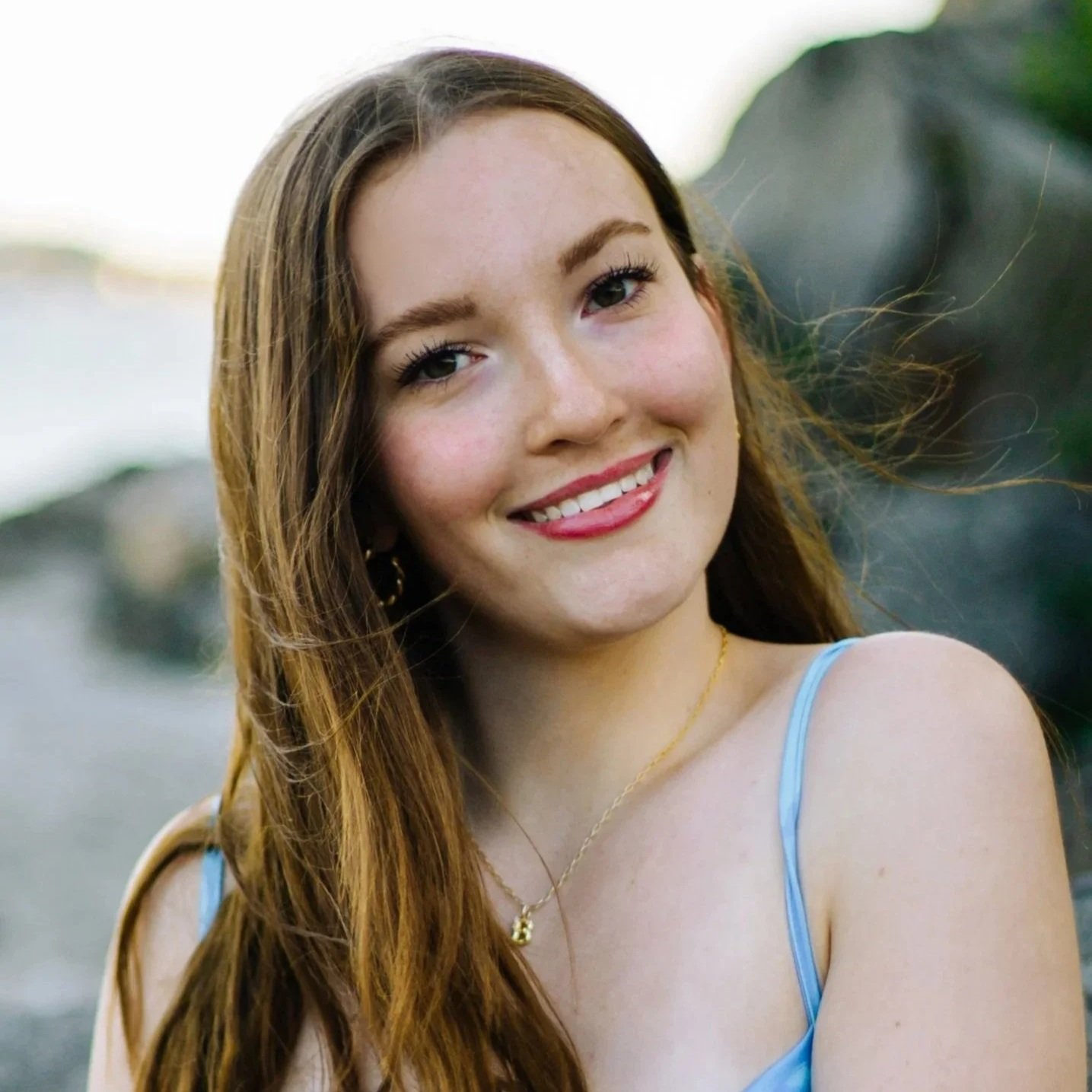Close-up of a young woman with long red hair, smiling, wearing a blue spaghetti strap top, with a blurred outdoor background.