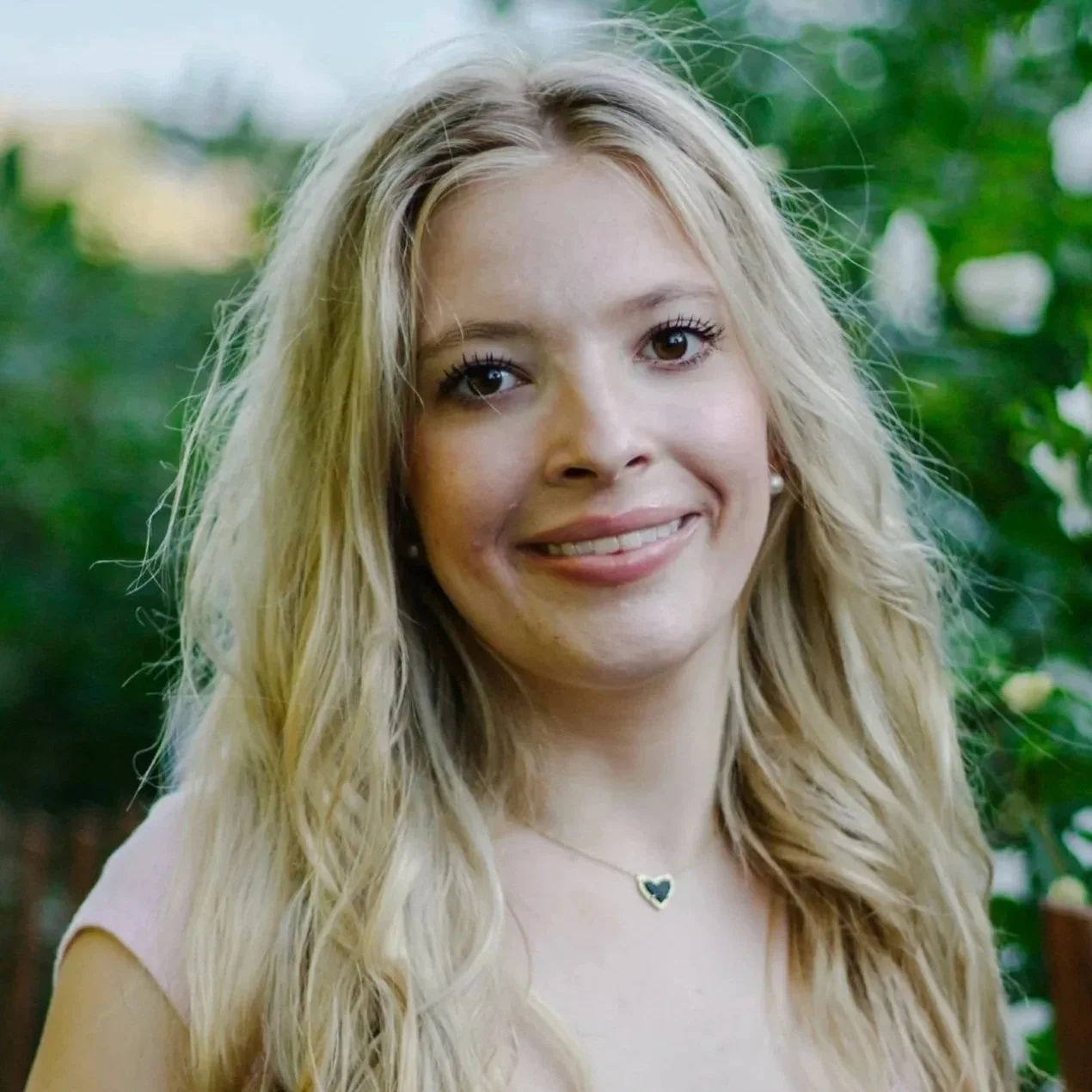 A young woman with long blonde wavy hair, smiling, wearing pearl earrings and a heart-shaped necklace, with green foliage in the background.