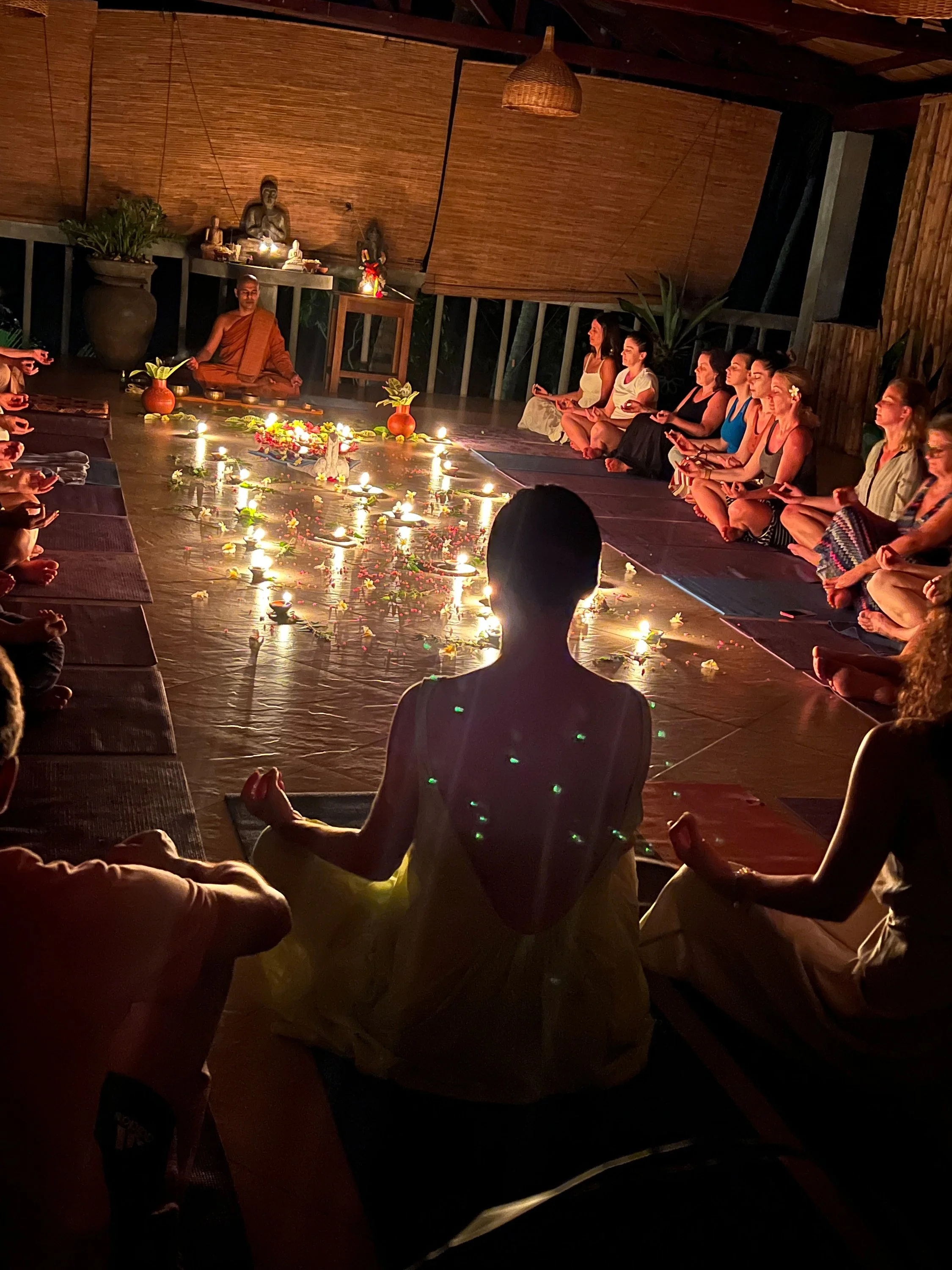Skupina lidí sedí se zkříženýma nohama kolem kruhu hořících svíček, účastní se meditace nebo duchovní ceremonie, vedené mnichem v místnosti zdobené soškami a rostlinami – inspirace pro ájurvédský wellness pobyt a meditativní zážitky.