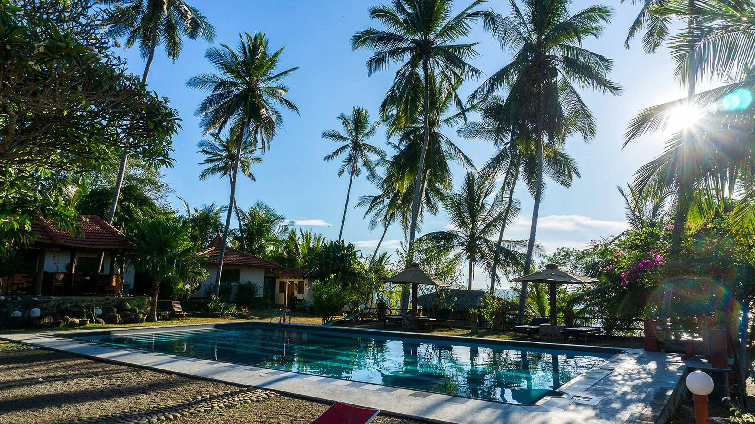Západ slunce nad tropickou pláží s palmami, loděmi u břehu a bujnou zelení – inspirace pro ájurvédský wellness pobyt a relaxaci u moře.