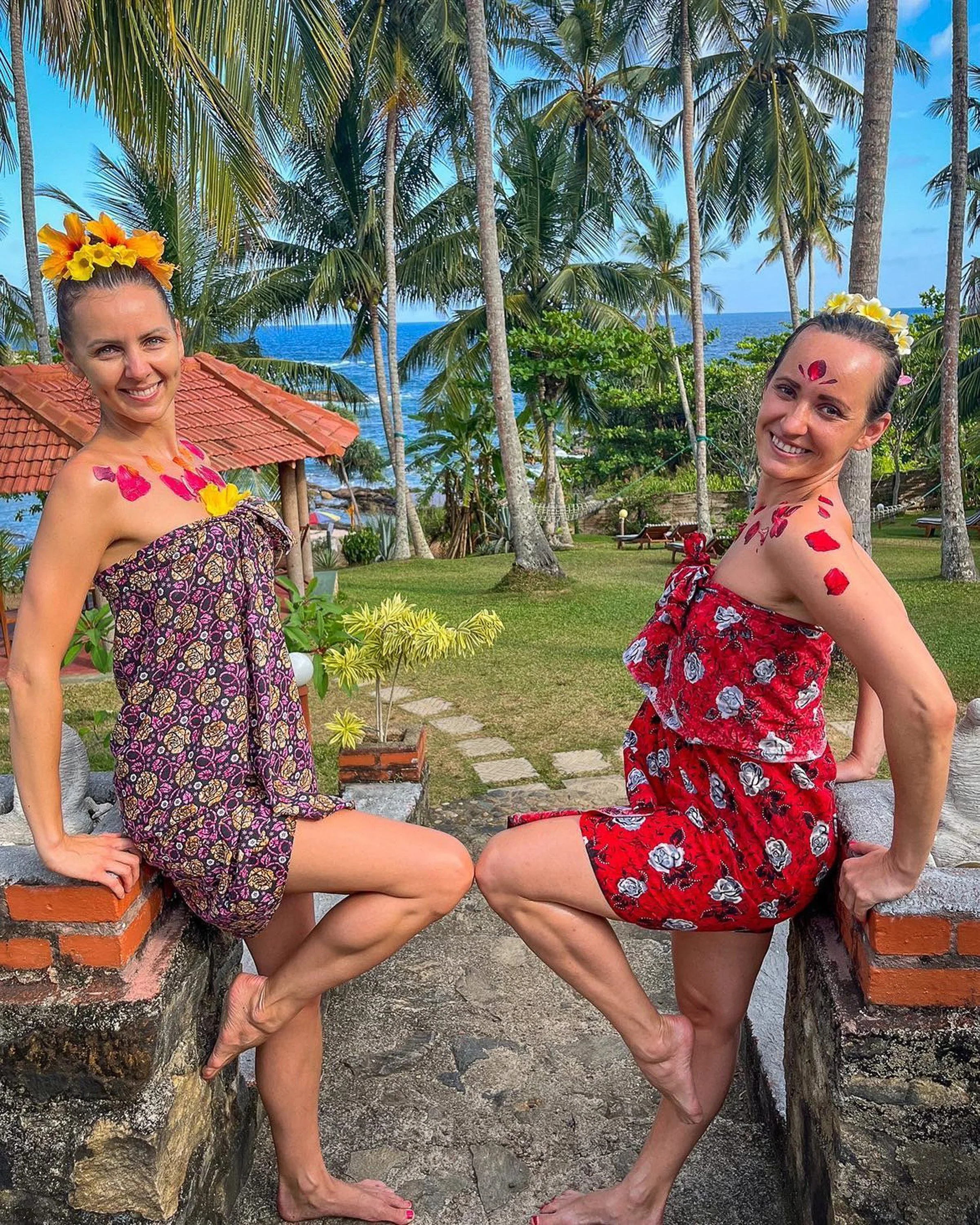 Dvě ženy v květinových ručnících s květinovými korunami a květinovou dekorací na obličeji a ramenou, sedící na cihlových a kamenných okrajích v tropické zahradě s palmami, trávou a výhledem na oceán, pod modrou oblohou – inspirace pro ájurvédský well