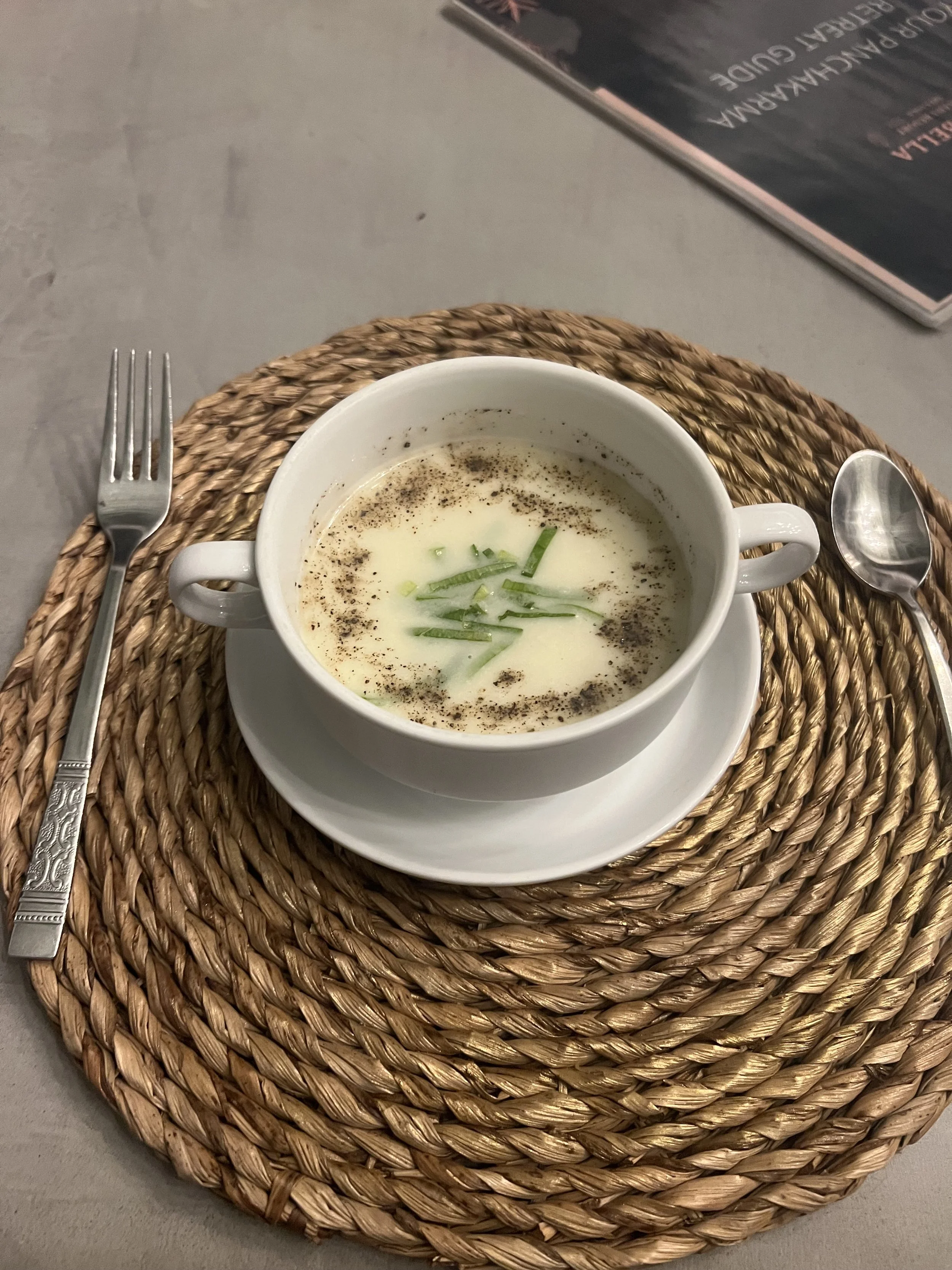 Miska krémové polévky s nasekanou jarní cibulkou na bílém talíři, na kulatém tkaném prostírání s vidličkou a lžící – inspirace pro ájurvédskou gastronomii a wellness pobyt.