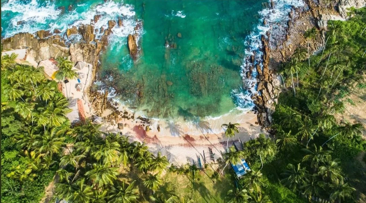 Letecký pohled na tropickou pláž s průzračně tyrkysovou vodou, obklopenou bujnými palmami a skalními útvary – inspirace pro ájurvédský wellness pobyt a relaxaci u moře.