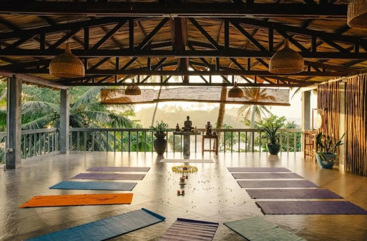 Open-air yoga studio s podložkami na dřevěné podlaze, obklopené bujnou zelení a tropickými stromy, se západem slunce v pozadí – inspirace pro ájurvédský wellness pobyt a relaxační cvičení jógy.