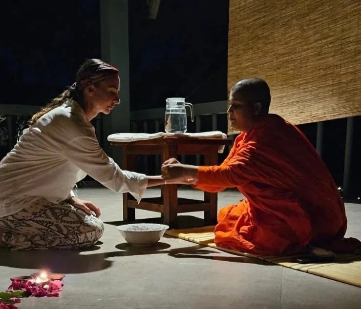 Žena a buddhistický mnich při duchovní výměně na dřevěné podlaze v noci, s miskou, stolkem a džbánem vody – inspirace pro ájurvédský wellness pobyt a duchovní zážitky.