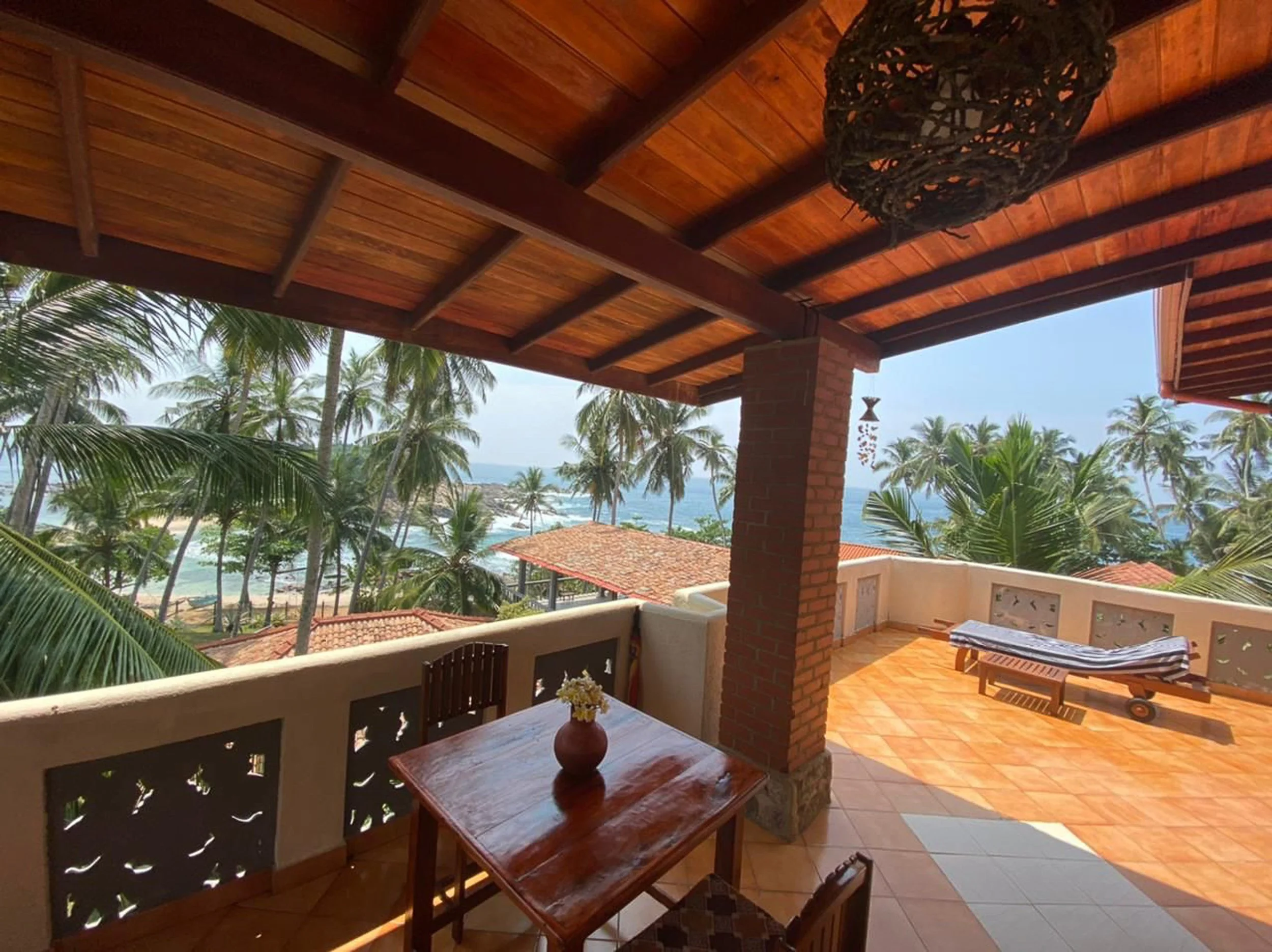 Výhled z balkonu na tropickou pláž s palmami, střechami s červenými taškami a oceánské vlny, balkon s dřevěným stolem, vázičkou s květinami, lehátkem a dekorací z mušlí – inspirace pro ájurvédský wellness pobyt a relaxaci u moře.