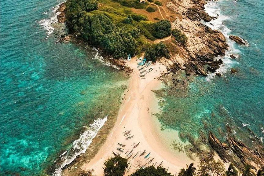 Aerial view of a small beach extending from a forested peninsula into the ocean, with boats and rocky surroundings.