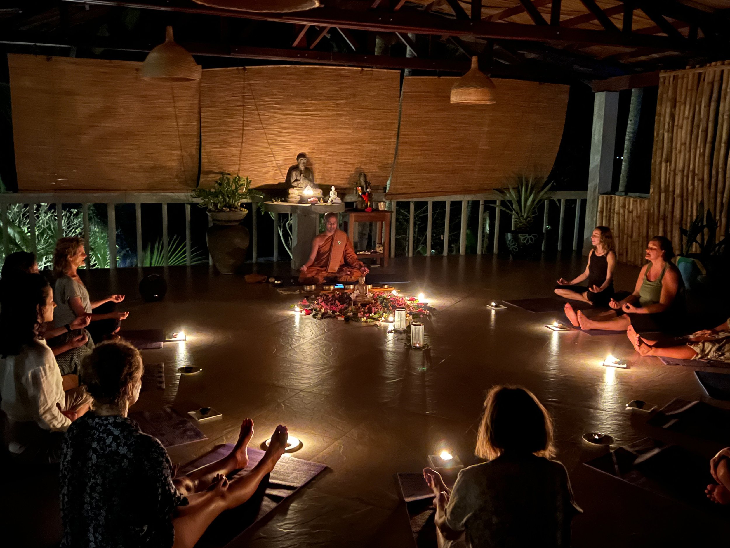 Lidé cvičí jógu v tlumeně osvětlené místnosti s hořícími svíčkami, rostlinami a soškou Buddhy vepředu – inspirace pro ájurvédský wellness pobyt a meditativní relaxaci.