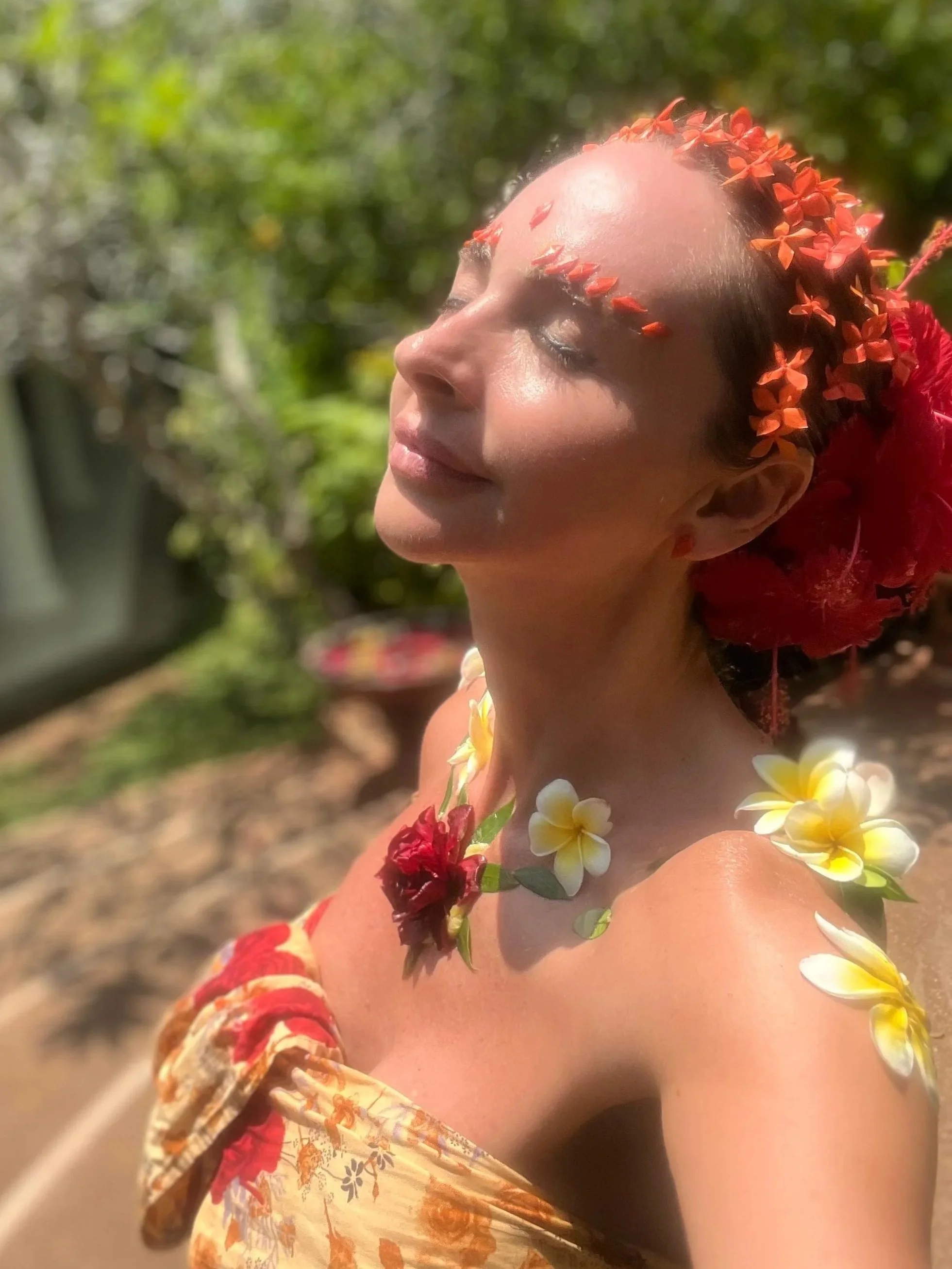 A woman with closed eyes and a peaceful expression, adorned with flowers on her forehead, neck, and shoulders, standing outdoors with greenery in the background.