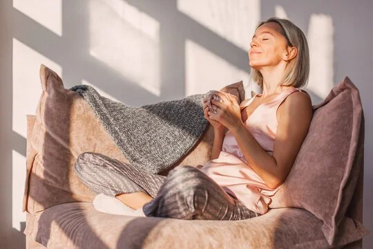 Žena sedí na posteli, usmívá se se zavřenýma očima a drží hrnek, sluneční světlo prosvítá do světlé místnosti.