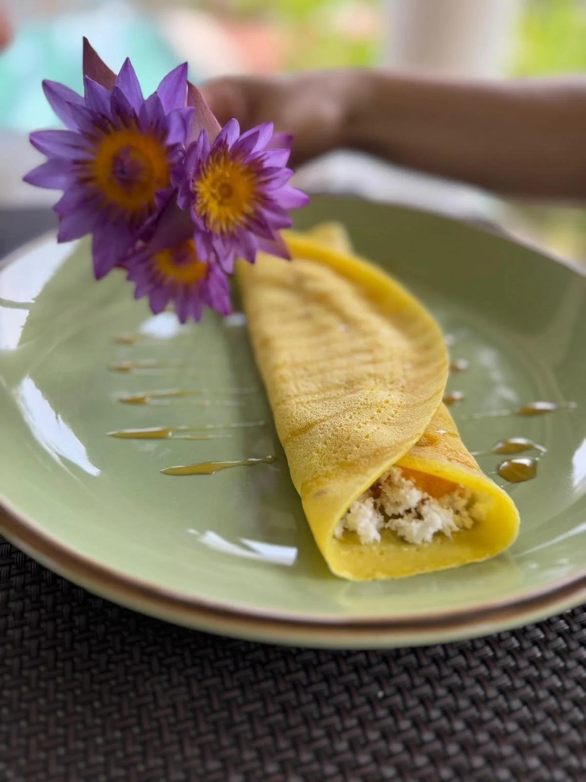 Žlutá rolovaná omeleta na zeleném talíři, ozdobená fialovými květy a politá sirupem, na tmavém tkaném prostírání – zdravá a estetická snídaně během ájurvédského wellness pobytu.