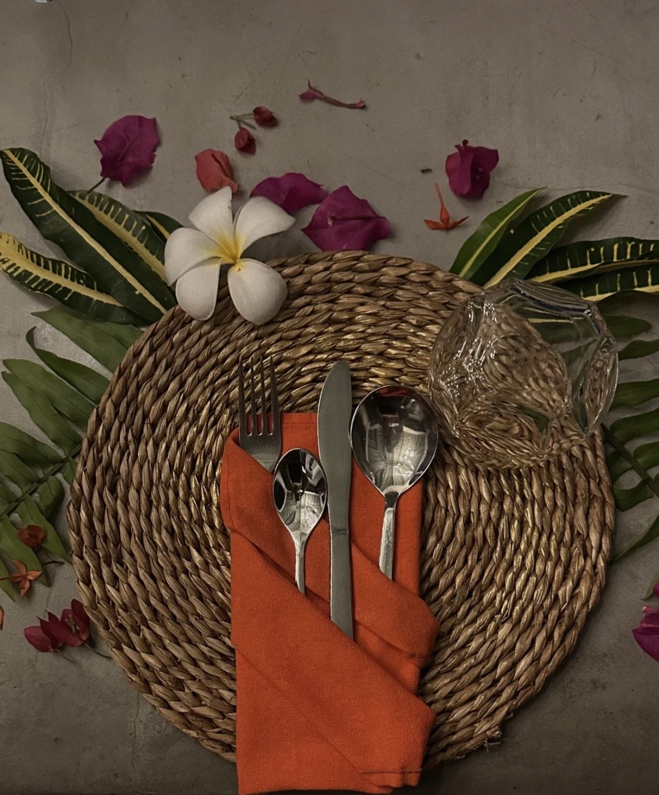 Stůl s tkaným prostíráním, oranžovým ubrouskem, příbory, prázdnou sklenicí a dekorací z listů a květin – inspirace pro ájurvédskou gastronomii a wellness pobyt.
