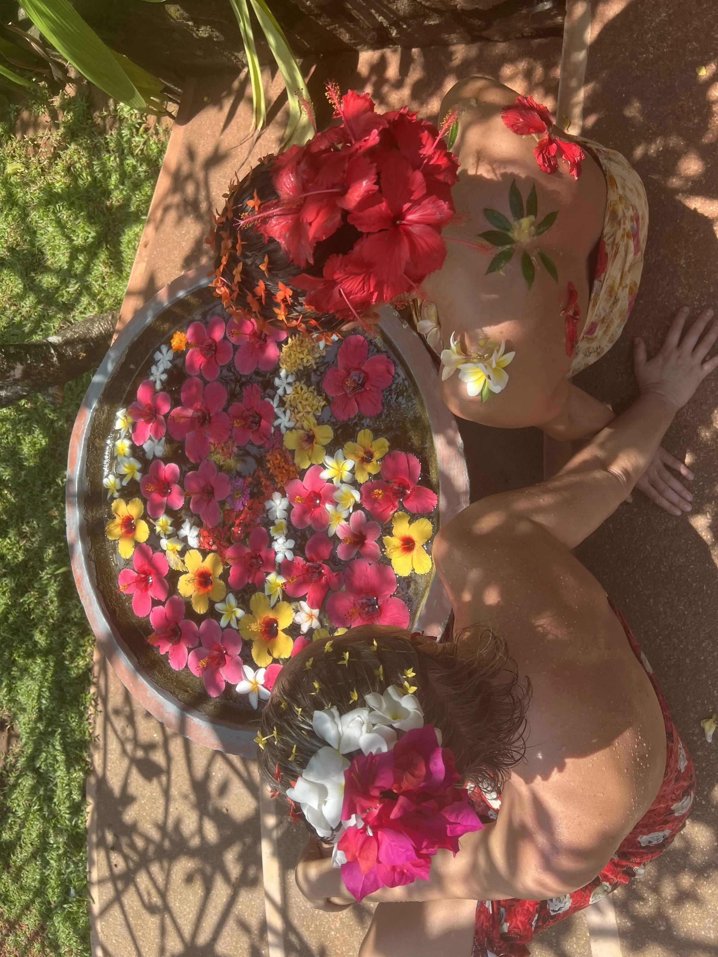 Dvě ženy s květinami ve vlasech koupe se ve vaně plné plovoucích barevných květin, venku s bujnou zelení a stíny – inspirace pro ájurvédský wellness pobyt a relaxaci v přírodě.