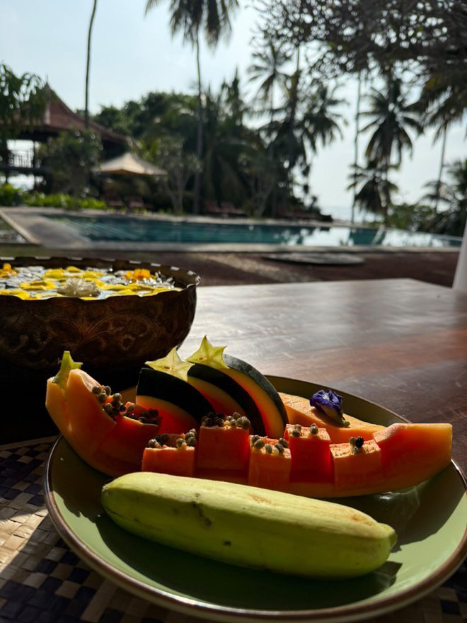 Plate of sliced papaya, melon, and watermelon with berries, in front of a pool surrounded by trees and tropical scenery.