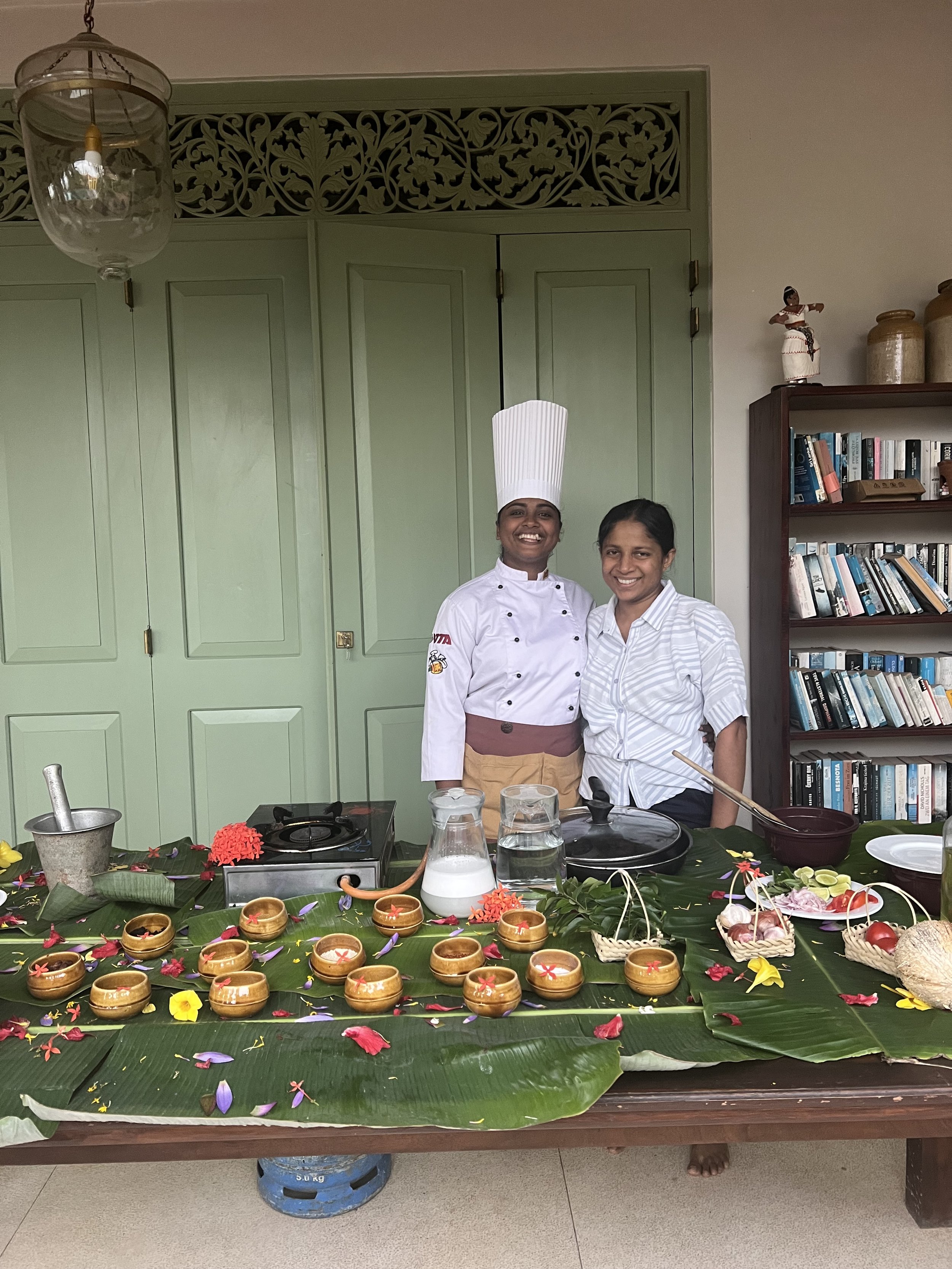 Dvě ženy stojí za stolkem s tradičním jídlem, květinami a listy, usmívají se do kamery – jedna v kuchařské uniformě, druhá v proužkované košili, ideální inspirace pro ájurvédskou gastronomii a wellness zážitky.