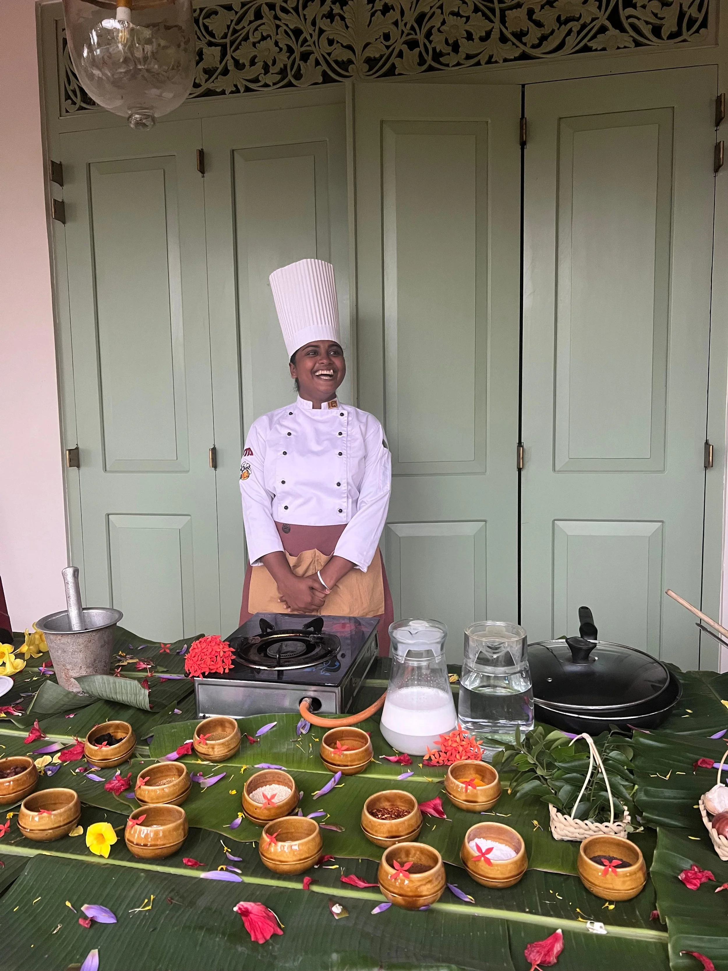 A smiling female chef in a white uniform and tall chef hat standing behind a table decorated with flower petals and banana leaves, with bowls, a mortar and pestle, a cooking stove, and pitchers of water and milk.