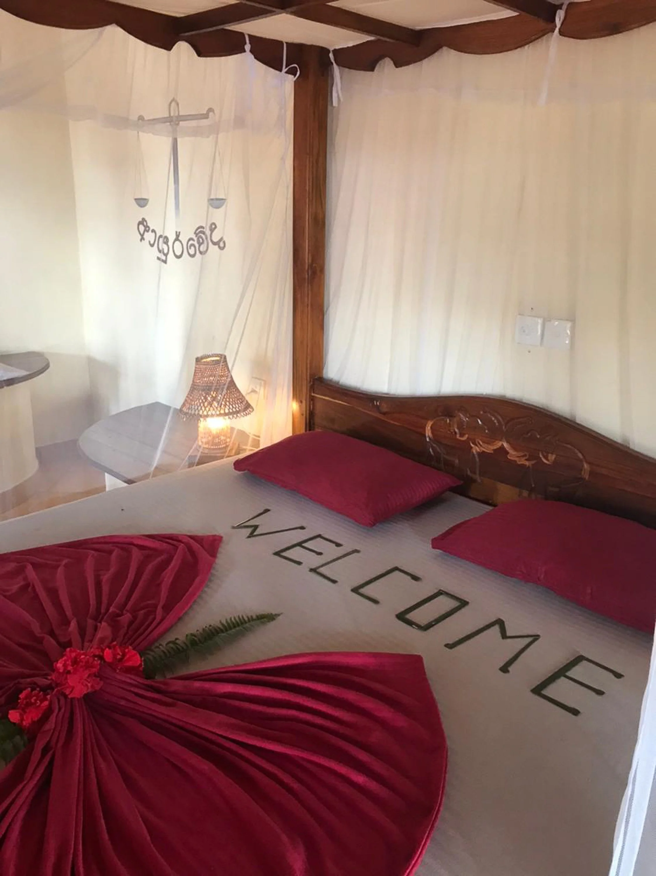 Hotelová postel s bílým prostěradlem s nápisem "WELCOME", dvěma růžovými polštáři, červeným ručníkem ve tvaru květu a malou kyticou červených květin, s moskytiérou, dřevěným čelem postele a lampou s proutěným stínidlem – inspirace pro ájurvédský well
