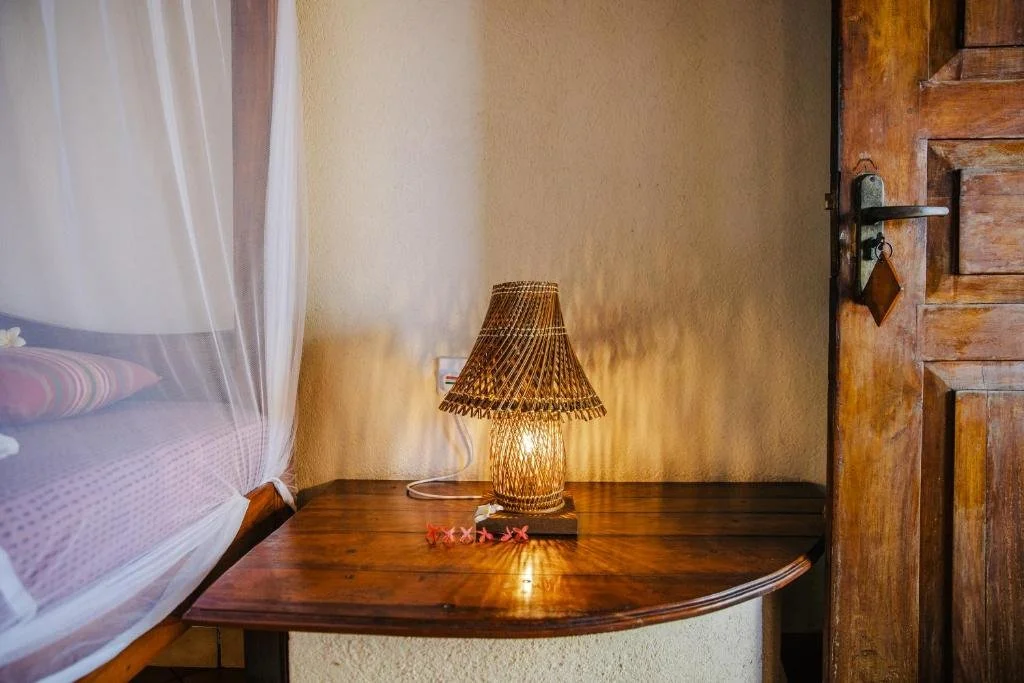 A wooden nightstand with a woven lamp emitting a warm glow. To the right is a closed wooden door with a latch. To the left, part of a bed with pink and purple bedding and a mosquito net canopy.