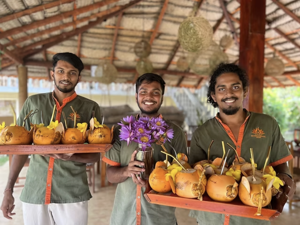 Three men standing in a wooden pavilion, each holding trays with coconuts and flowers, smiling at the camera.