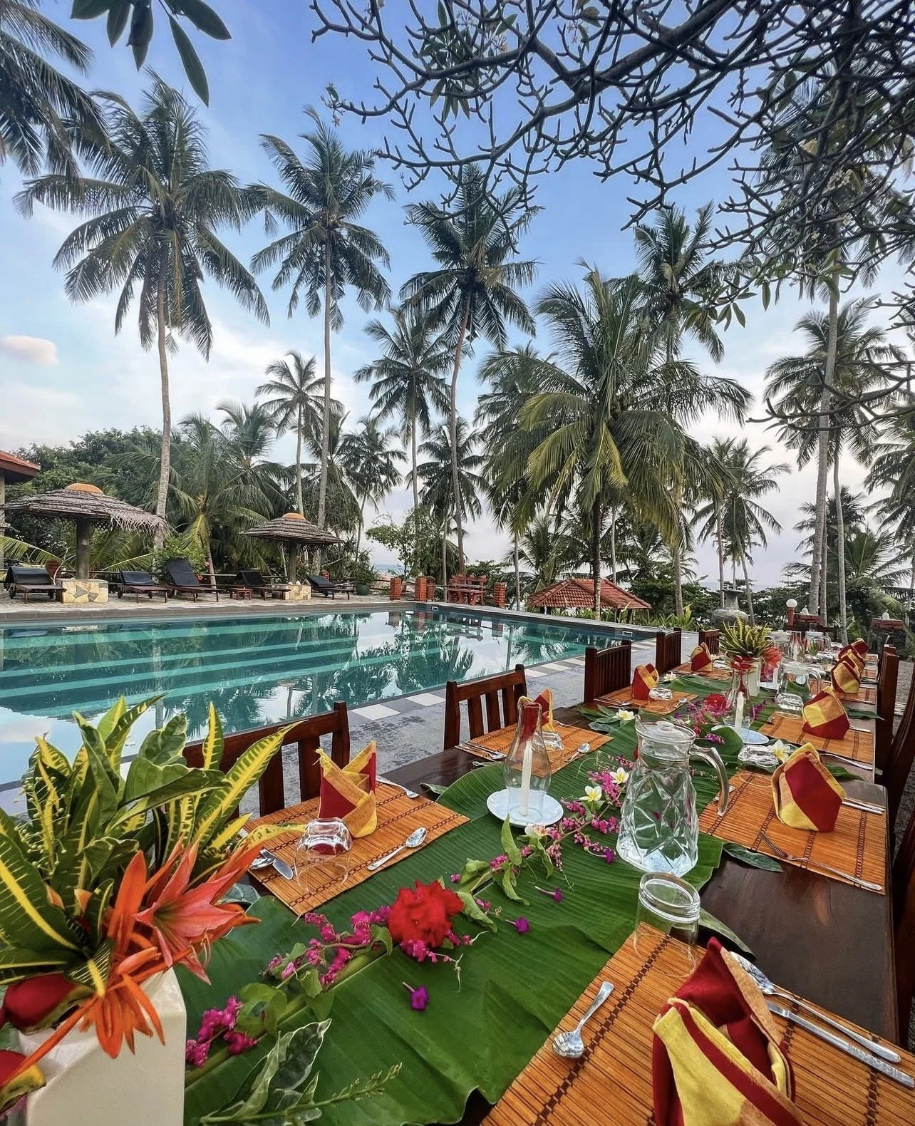 Tropické venkovní stolování u bazénu s dlouhým stolem, barevnými ubrousky, květinami a sklenicemi, obklopené palmami – inspirace pro ájurvédský wellness pobyt a exotické zážitky.
