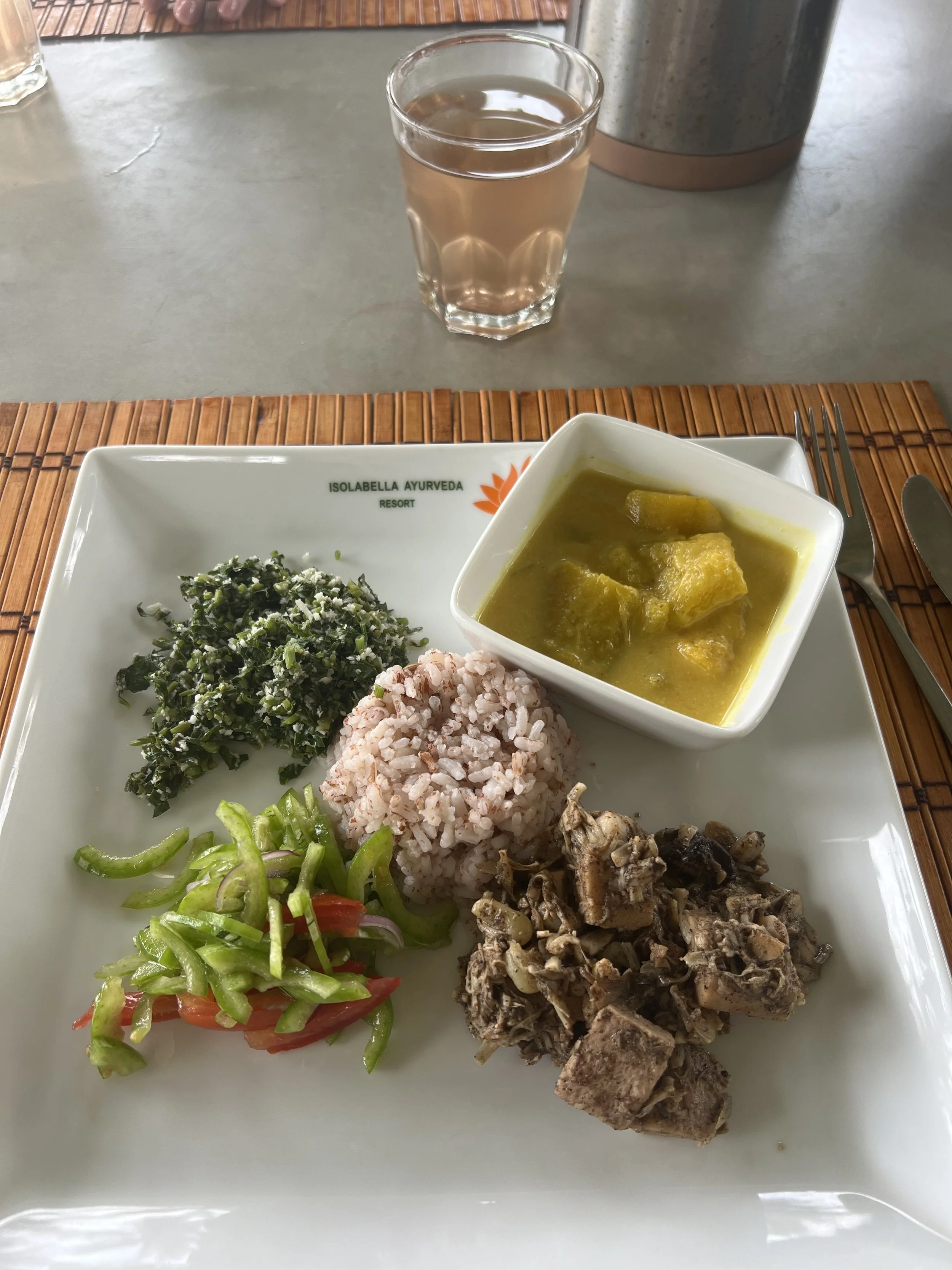 Talíř indického vegetariánského jídla s rýží, restovanou zeleninou, zeleninovým kari a směsí zeleniny, s sklenicí vody na bambusovém prostírání – inspirace pro ájurvédskou gastronomii a wellness pobyt.