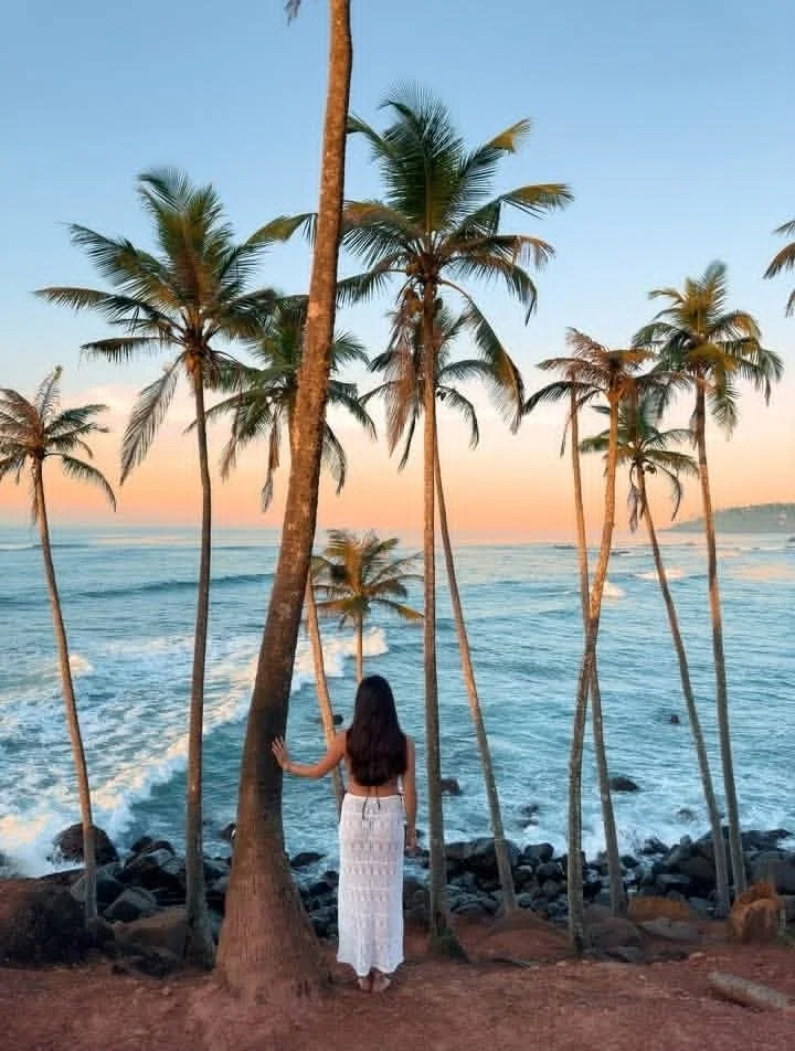 Žena s dlouhými tmavými vlasy v bílé háčkované šatičce stojí na skalnaté pláži, drží palmu, s vlnami oceánu a západem slunce v pozadí – inspirace pro ájurvédský wellness pobyt a relaxaci u moře.