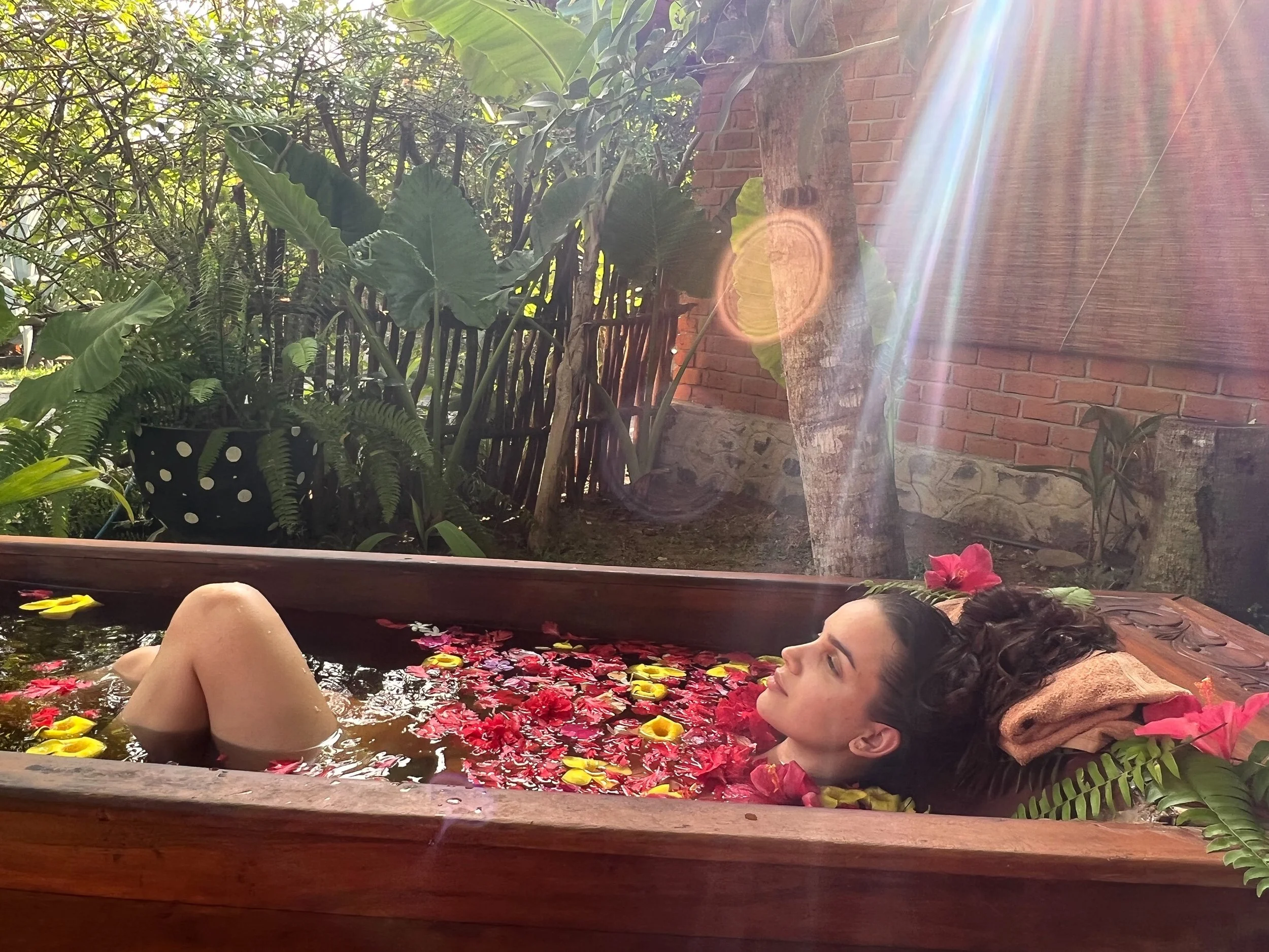 Žena relaxuje v dřevěné vaně naplněné vodou a květinovými okvětními lístky, venku mezi zelenými rostlinami a slunečním světlem.