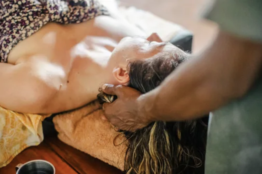 A woman lying face-up on a massage table, receiving a massage from a person with darker skin. The woman has brown hair with blonde highlights and appears relaxed.